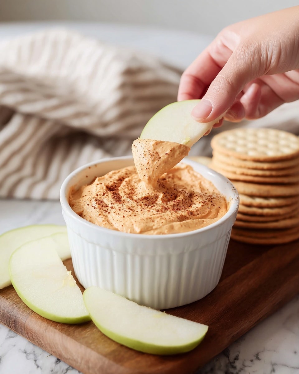 A white ramekin filled with a thick, creamy light orange dip topped with a sprinkle of dark brown spice is centered on a wooden board with a few slices of pale green apple around it. A woman's hand holds one pale green apple slice dipped halfway into the smooth, slightly textured dip, showing the contrast between the fresh fruit and the spread. In the background, a soft striped cloth and stacked crackers create a cozy setting on a white marbled surface. photo taken with an iphone --ar 4:5 --v 7