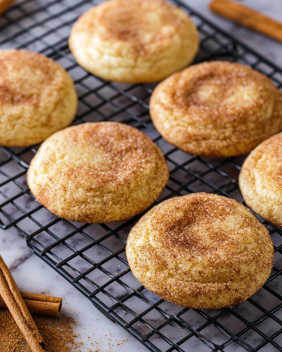 The image shows six round cookies with a light golden brown color, covered with a dusting of cinnamon and sugar. The cookies have slightly wrinkled tops and soft edges, arranged in two rows on a black cooling rack. The rack sits on a white marbled surface, and there are two cinnamon sticks and some loose cinnamon powder visible near the corner. The lighting highlights the texture of the cinnamon sugar coating and the warm, soft look of the cookies. photo taken with an iphone --ar 4:5 --v 7
