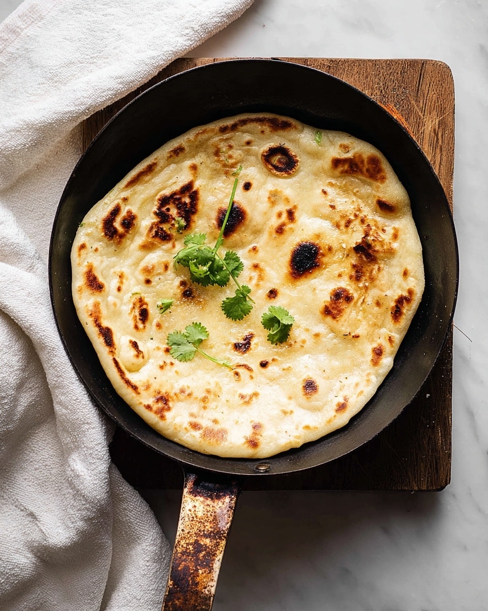 A single round flatbread with a golden-brown surface and several darker toasted spots lies in a black frying pan. The flatbread’s texture is soft and slightly puffy, with small bubbles scattered across. Three small sprigs of fresh green cilantro rest near the center, adding a bright contrast. The pan handle shows a rustic patina, and the pan sits on a piece of worn wooden board above a white marbled surface. A white cloth is casually placed to the left side. Photo taken with an iphone --ar 4:5 --v 7