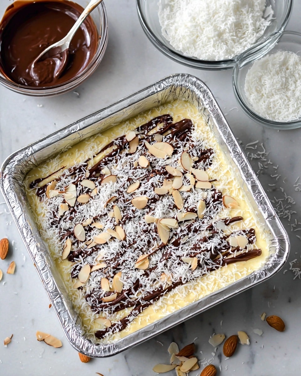 A square glass baking dish lined with shiny silver foil holds a three-layer dessert. The bottom layer is pale yellow, smooth, and creamy. On top, there is a thin drizzle of dark brown chocolate spread unevenly over the surface. The dessert is sprinkled with white shredded coconut and light beige slivered almonds scattered loosely all over. Around the baking dish, there are some extra almond slivers scattered on the white marbled surface. In the background, there are two clear glass bowls, one filled with white shredded coconut and the other with chocolate sauce, with a silver spoon resting inside the chocolate bowl. photo taken with an iphone --ar 4:5 --v 7