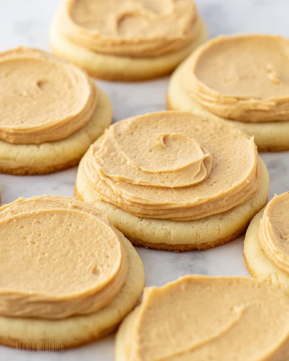 This image shows several round sugar cookies with a thick layer of light brown peanut butter frosting spread smoothly on top. Each cookie has two layers: the bottom layer is a pale yellow cookie, soft and smooth, and the top layer is a creamy, slightly textured peanut butter frosting, evenly covering the cookie surface but not going to the edges. The cookies are arranged close together on a white marbled surface. The lighting is bright and natural, showing the soft texture of both the cookie and frosting clearly. photo taken with an iphone --ar 4:5 --v 7