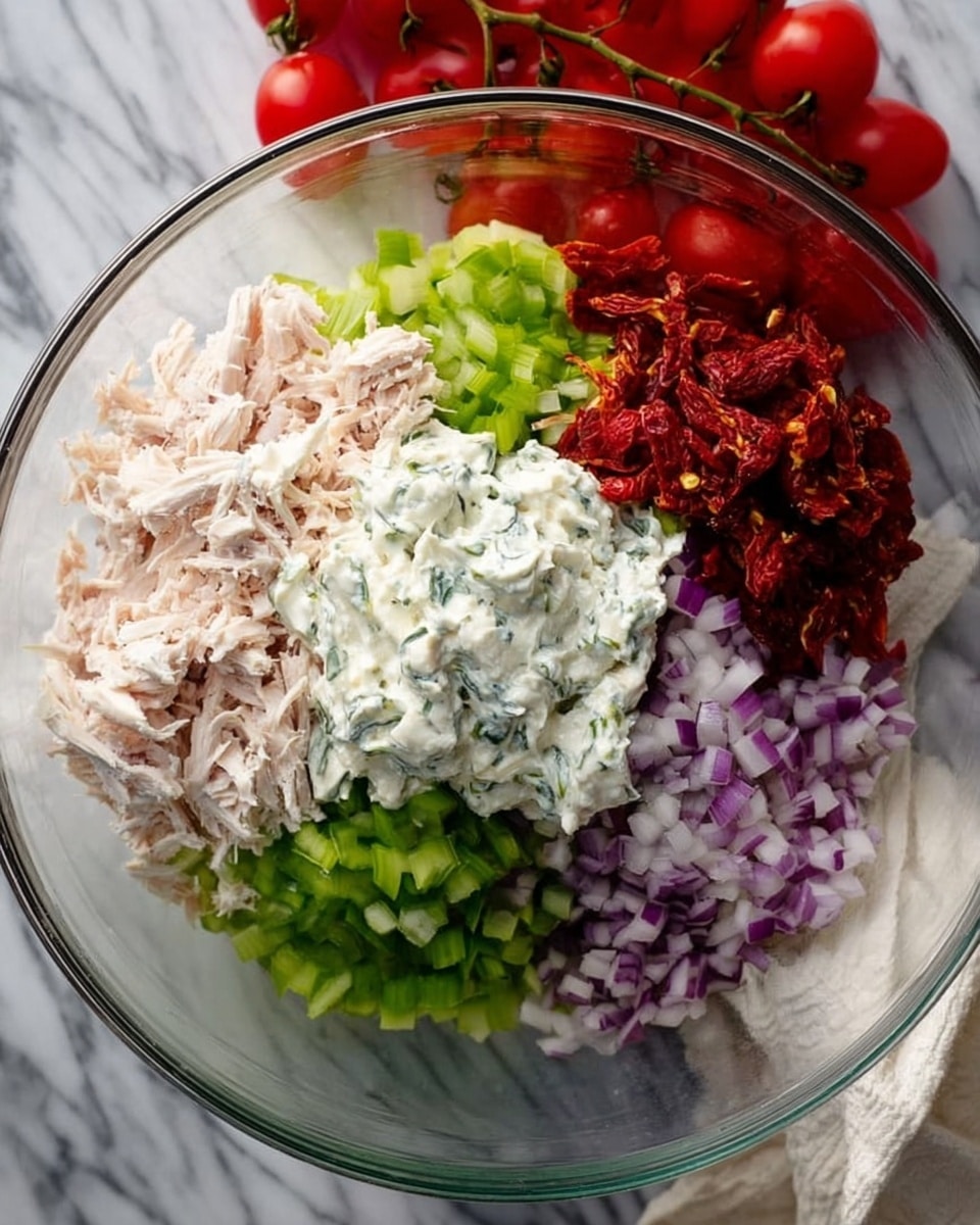 A clear glass bowl on a white marbled surface holds five colorful layers of ingredients arranged separately. Starting from the top left is shredded pale pink cooked chicken, to the right is a pile of dark red diced sun-dried tomatoes, below that is a heap of bright green chopped celery, next to it on the left is finely chopped purple onions, and in the center is a dollop of white creamy sauce with green herb bits mixed in. A bunch of fresh red cherry tomatoes are blurred in the background above the bowl. A woman's hand is holding a white cloth near the bottom right side of the bowl. Photo taken with an iphone --ar 4:5 --v 7