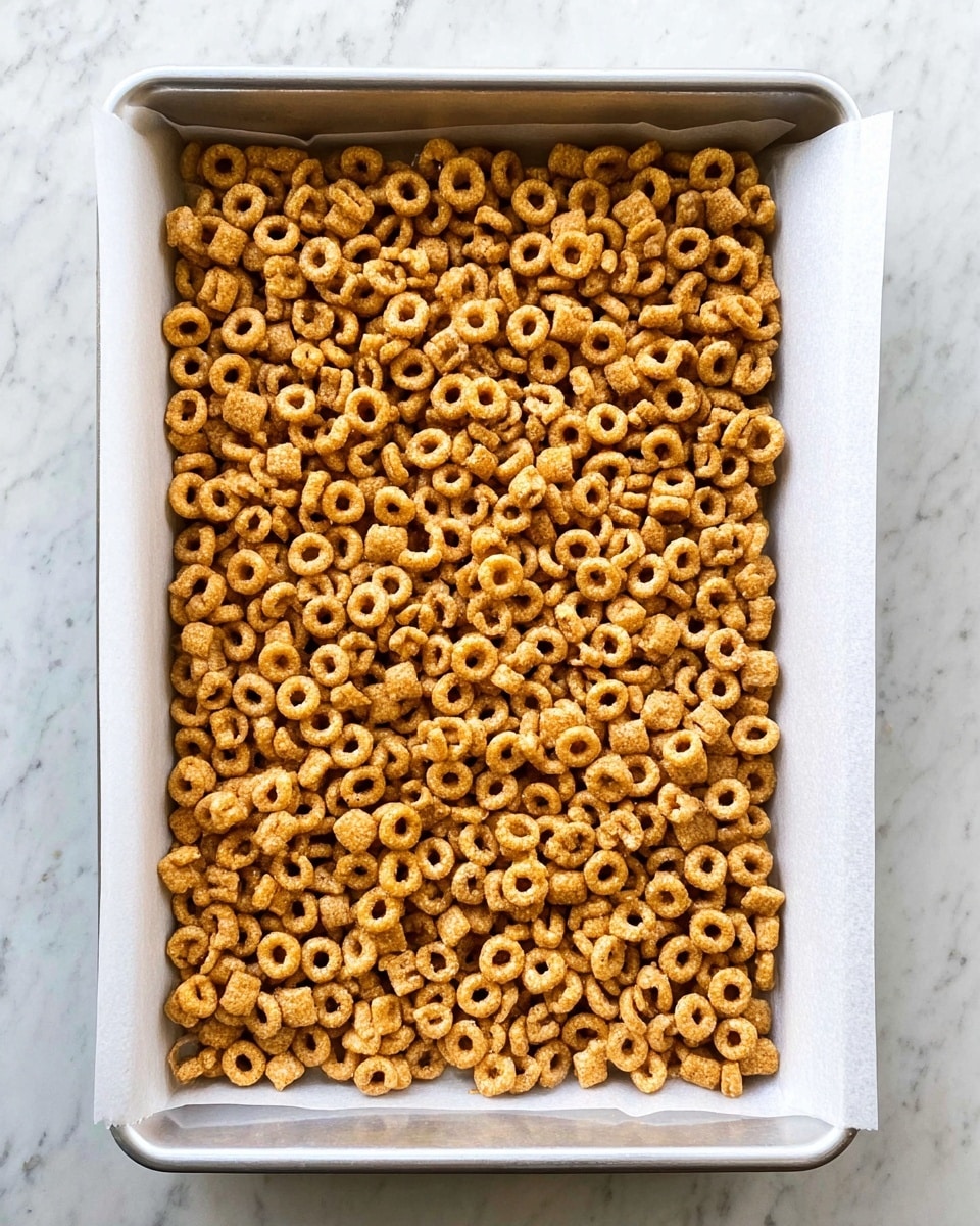 A rectangular baking tray lined with white parchment paper holds a thick, even layer of small, round cereal pieces that are golden brown in color. The cereal pieces have a slightly rough texture and fill the entire tray, creating a dense, uniform surface. The tray sits on a white marbled surface, adding a clean and bright background to the scene. Photo taken with an iphone --ar 4:5 --v 7