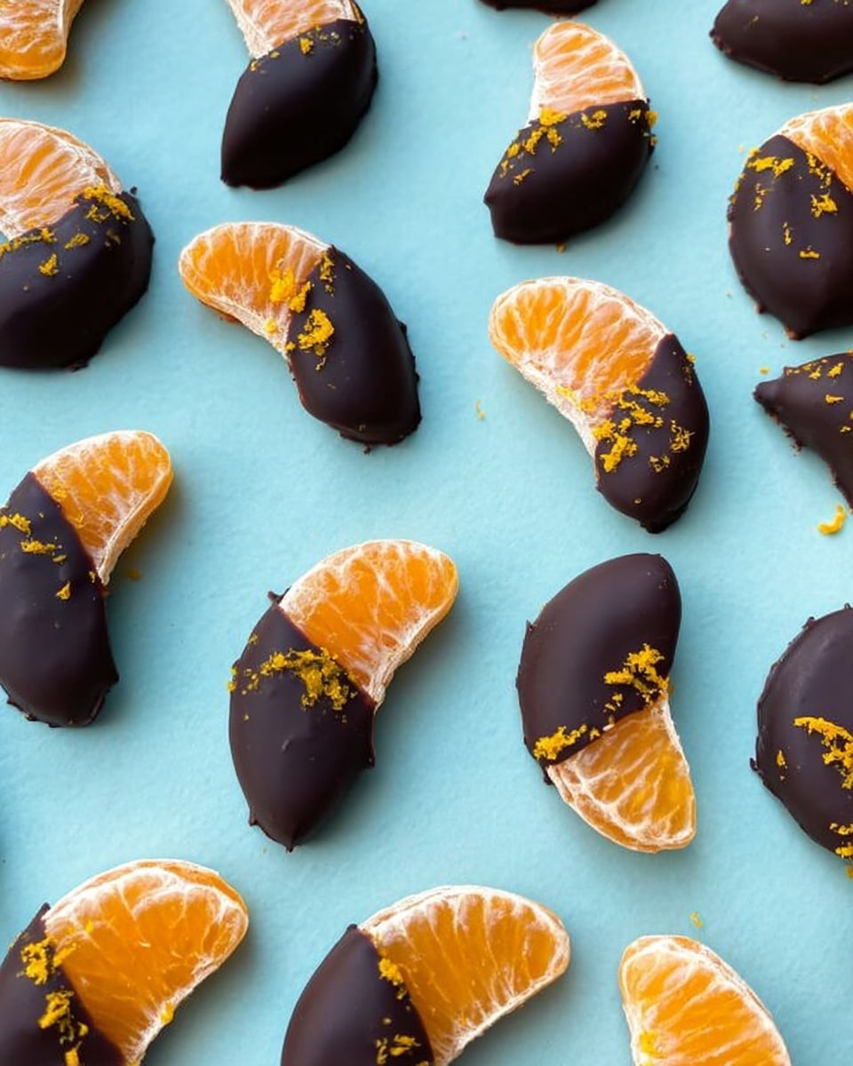 The image shows many small orange tangerine slices, each half covered in smooth dark chocolate, laid out evenly on a pale blue surface. Each slice has a bright orange zest sprinkled on top, adding fine texture and contrast. The tangerine segments have a juicy, slightly translucent look with clear segments visible, while the chocolate coating is glossy and thick, covering about half of each slice’s curved side. The arrangement is casual but spaced out, showing the two different textures and colors side by side. Photo taken with an iphone --ar 4:5 --v 7