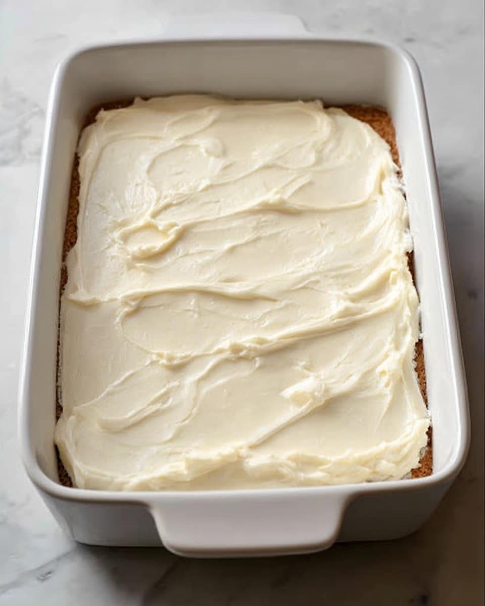 A white rectangular baking dish contains a dessert with two visible layers: the bottom layer is a pale brown cake or crust, and it is topped with a thick, creamy, smooth white frosting evenly spread across the surface. The texture of the frosting is soft and lightly swirled, covering the entire top of the dessert. The dish is placed on a white marbled texture surface. photo taken with an iphone --ar 4:5 --v 7