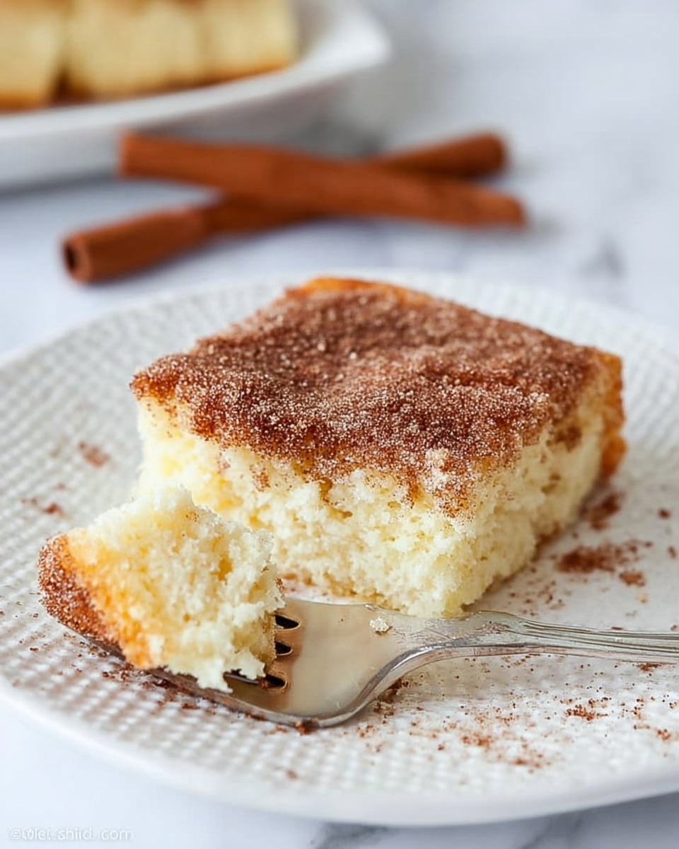 The image shows a square slice of soft, light yellow cake with a thick topping layer of brown cinnamon sugar sprinkled on top. A small piece of the cake is held on a silver fork, showing the contrast between the pale inside of the cake and the darker, crunchy topping on the outside. The cake sits on a white textured plate, next to a whole cinnamon stick, with some cinnamon sugar crumbs scattered around. The background is a white marbled surface. photo taken with an iphone --ar 4:5 --v 7