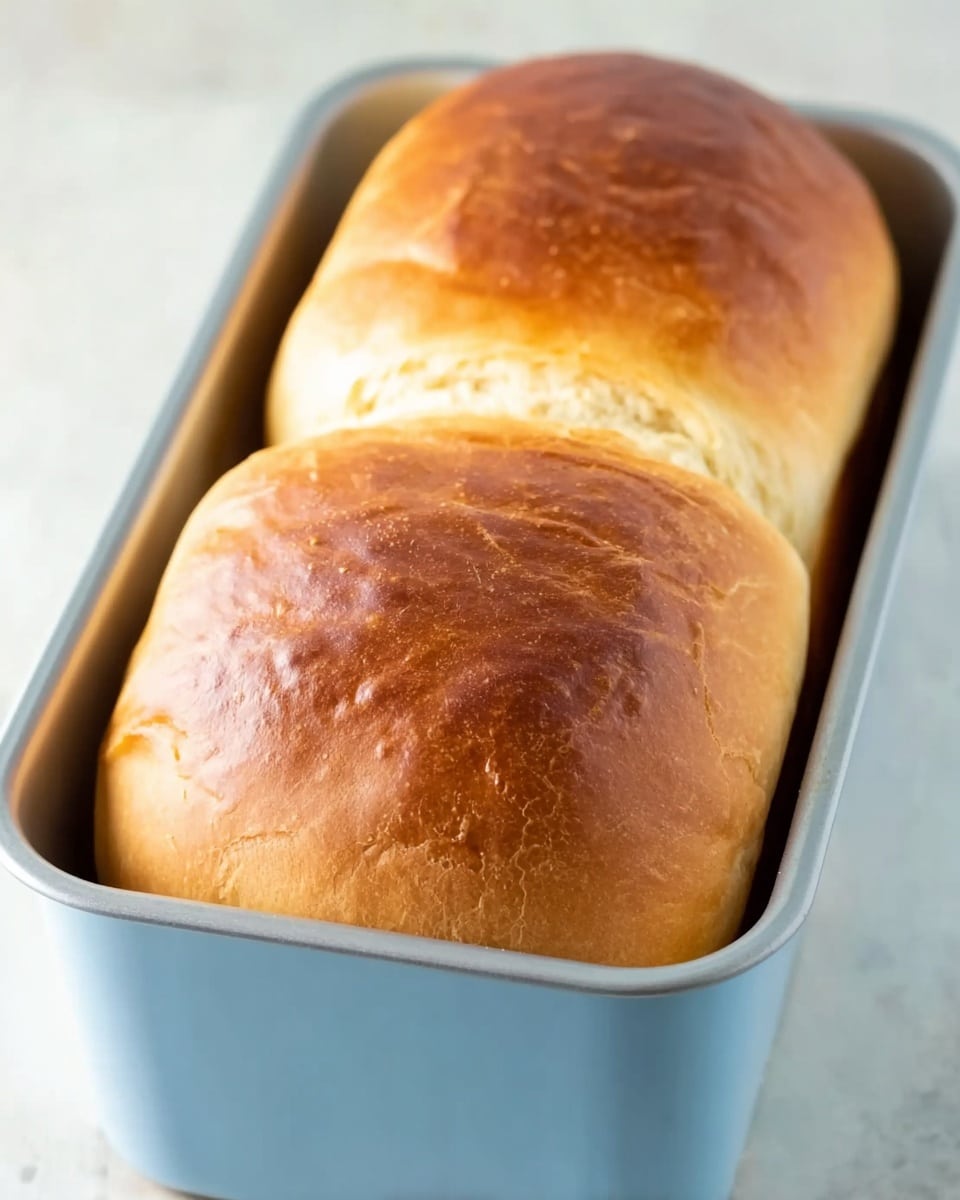 The image shows two soft, golden brown bread loaves rising side by side in a light blue loaf pan. The tops of the bread are smooth with a few small cracks, displaying different shades of brown, from light golden to a darker toast color at the front loaf. The loaf pan sits on a white marbled surface, adding a clean and bright feel to the image. The bread looks fresh, with a slightly shiny crust but no crumbs or extra toppings visible. Photo taken with an iphone --ar 4:5 --v 7