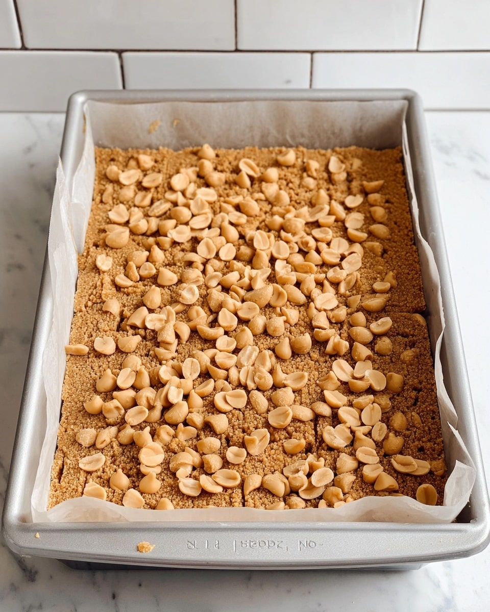 The image shows a rectangular baking pan lined with parchment paper, filled with one layer of light brown crumbly base that looks like crushed graham crackers or a similar crumb mixture. On top, there is an even scatter of tan-colored peanut butter chips and salted peanuts spread across the entire surface, giving a mix of smooth and rough texture. The pan and parchment paper edges are clearly visible, the pan is silver, and the setup is placed on a white marbled surface with a white tiled wall in the background. photo taken with an iphone --ar 4:5 --v 7