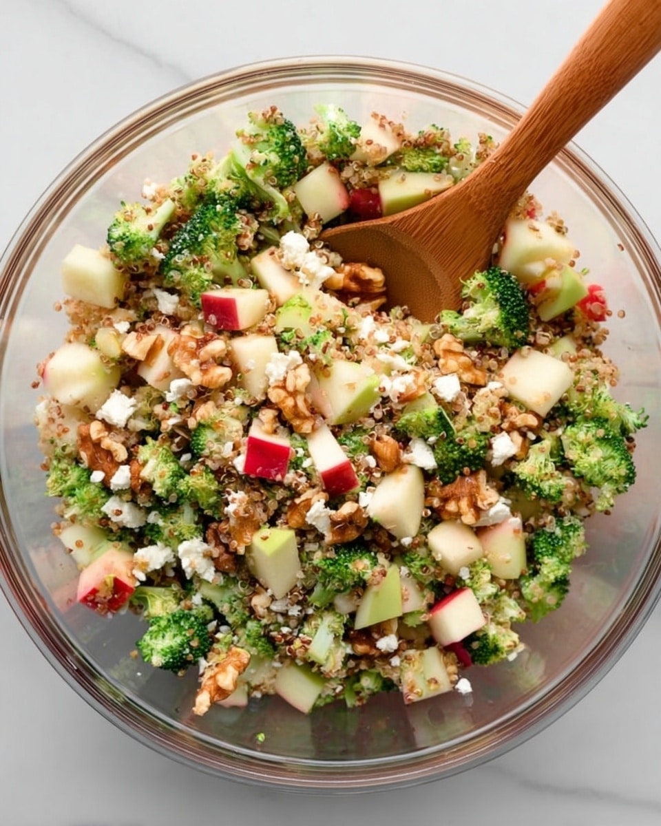 A clear glass bowl filled with a colorful salad featuring a mix of small, green broccoli florets, diced red and white apples, crumbled white cheese, light brown walnuts, and light tan quinoa all mixed together. A wooden spoon is placed inside the bowl as if ready to scoop up the salad. The bowl sits on a white marbled surface. photo taken with an iphone --ar 4:5 --v 7