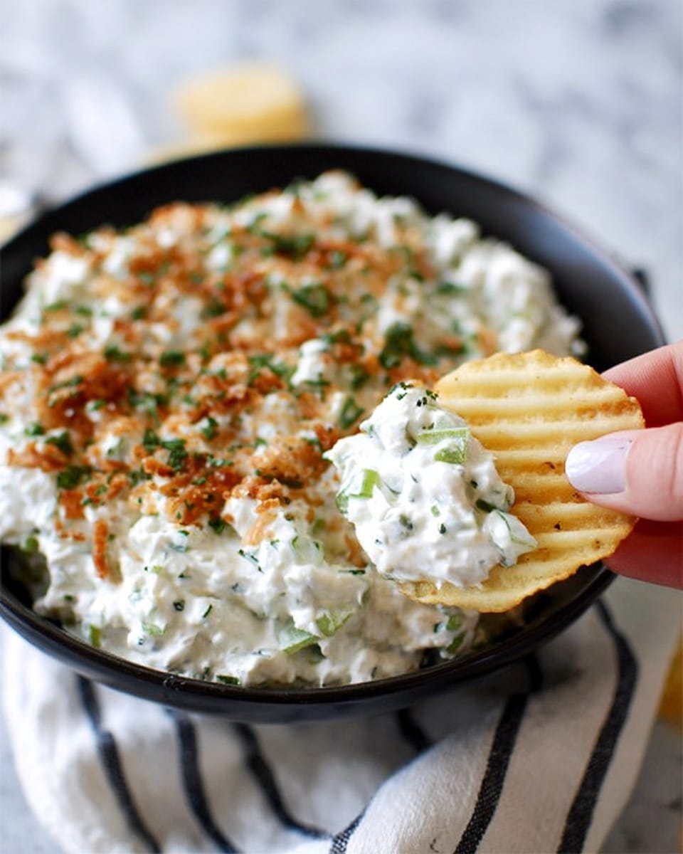 The image shows a close-up of a black bowl filled with creamy white dip that has green herb pieces and small brown crispy bits sprinkled on top. In the foreground, a woman's hand is holding a crinkled ridged potato chip covered with the same dip. The background surface is a white marbled texture with a white cloth featuring thin black stripes visible near the bowl. The dip has a thick, soft texture with small green herb bits mixed throughout and sprinkled on top, adding a fresh look. Photo taken with an iphone --ar 4:5 --v 7