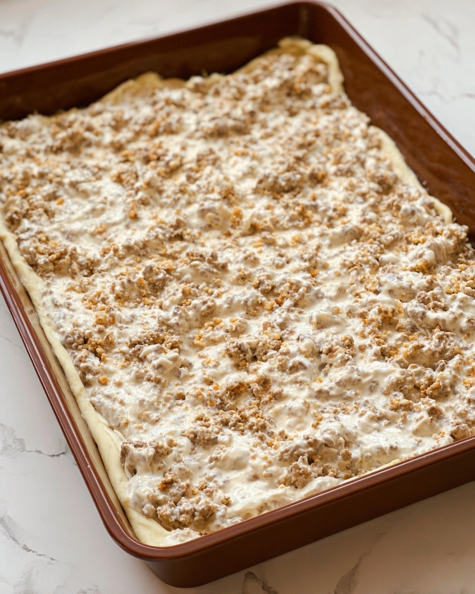 The image shows a rectangular baking dish filled with an uncooked layered dish. The bottom layer is a smooth dough crust pushed up slightly along the edges. On top of the crust is a thick, uneven layer of cooked ground meat mixed with a creamy white sauce, creating a textured and slightly chunky surface with various light beige and off-white tones. The baking dish is brown, placed on a white marbled surface. photo taken with an iphone --ar 4:5 --v 7