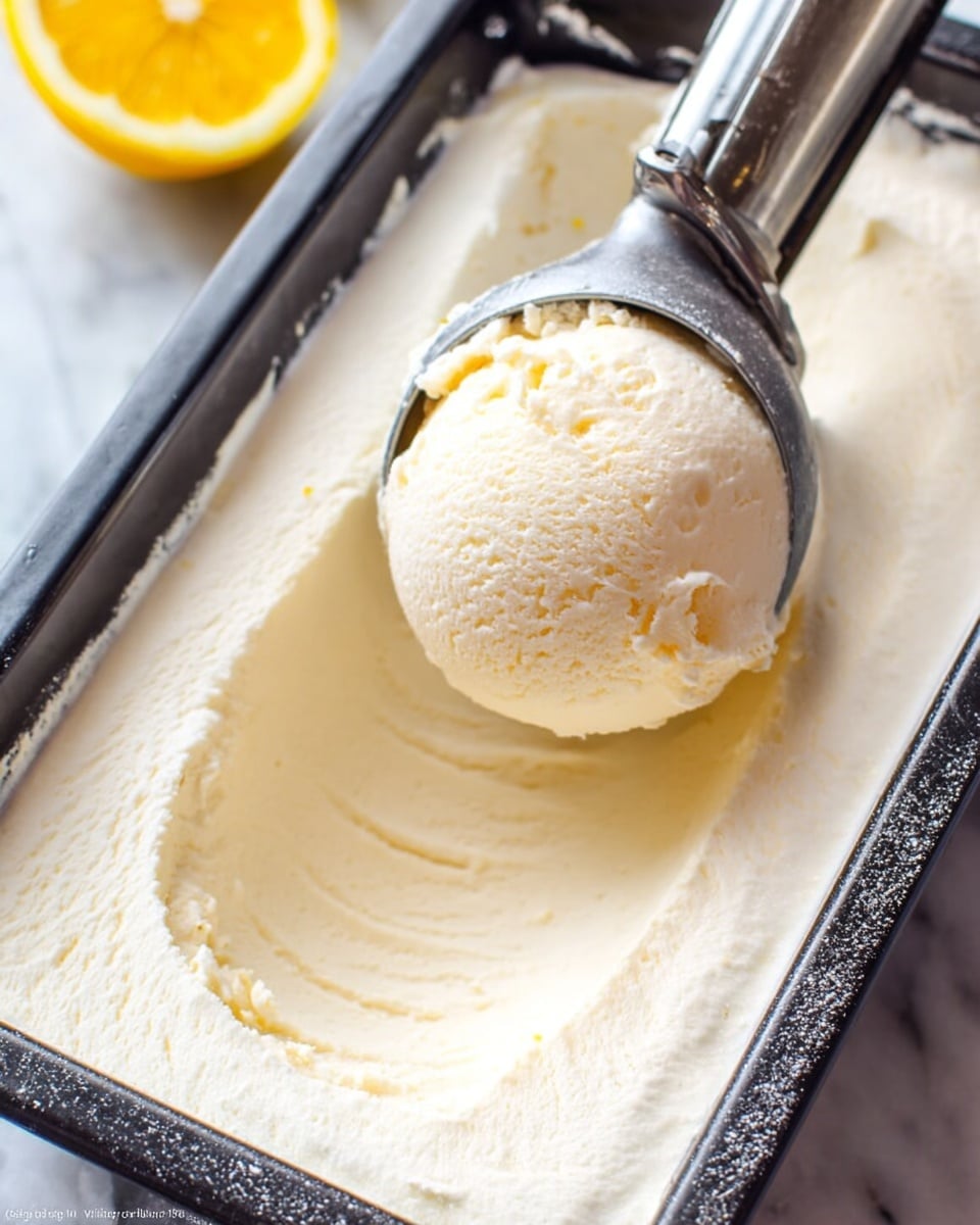 A close-up view of one scoop of light cream-colored ice cream being lifted with a silver ice cream scoop from a black rectangular container filled with creamy, smooth ice cream. The ice cream has a soft texture with slight ridges where it has been scooped, and the edges of the container show a fine layer of frost. A blurred yellow lemon half is visible in the background on a white marbled surface photo taken with an iphone --ar 4:5 --v 7