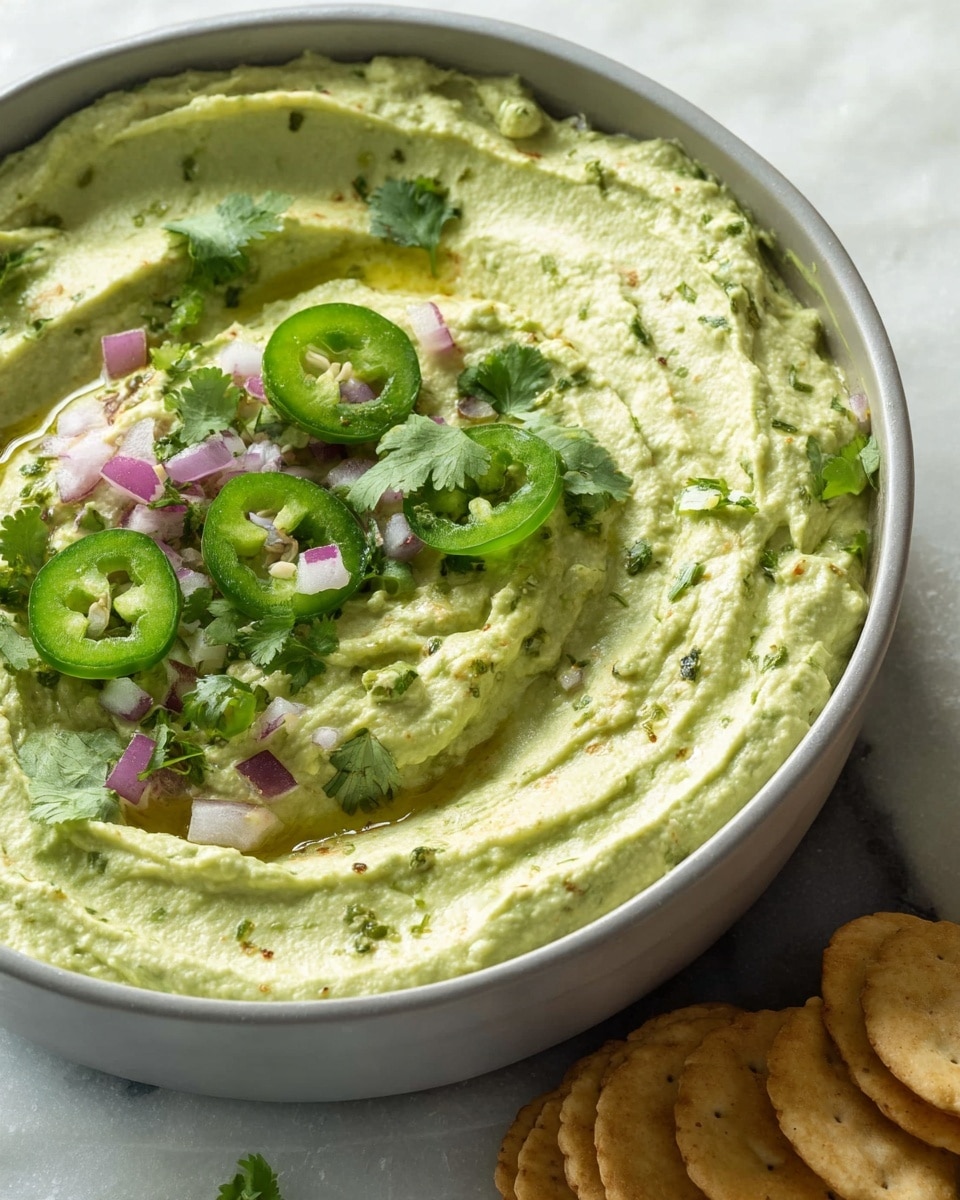 A bowl filled with a thick creamy green dip, having visible soft swirls on the surface. The dip is topped with thin slices of green jalapeño, small pieces of chopped purple onion, and fresh cilantro leaves scattered in various spots. There are small pools of oil in some dips’ crevices, adding shine and depth. The bowl is white, and the setting is a white marbled texture. A small stack of round crackers appears at the bottom right corner of the image photo taken with an iphone --ar 4:5 --v 7