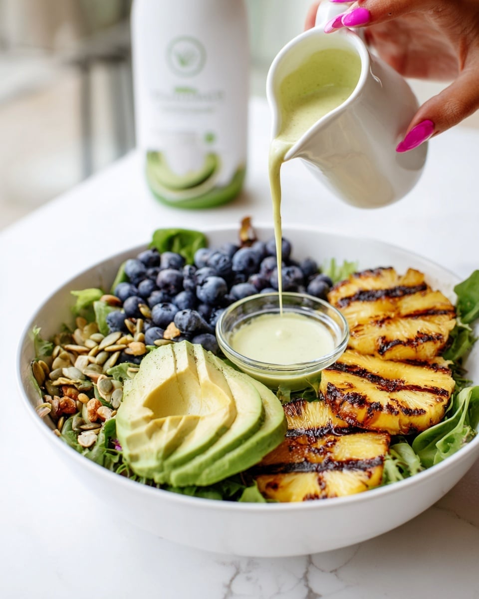 The image shows a white bowl filled with a fresh salad on a white marbled surface. The salad has a base layer of mixed green leaves. On top, there are three grilled pineapple slices on the right side, a half avocado sliced thinly with green flesh in the middle, and a cluster of fresh blueberries on the left. Scattered around are pumpkin seeds and bits of nuts for crunch. A small glass bowl with a pale green dressing sits inside the bowl near the blueberries. A woman's hand with pink nail polish is pouring creamy, light green dressing from a white pitcher over the avocado. In the background, a white bottle with green label details is slightly blurred. photo taken with an iphone --ar 4:5 --v 7