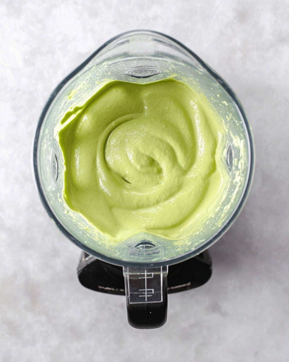 A clear blender jar seen from above filled with a smooth, bright green sauce that has a creamy texture with gentle swirls on the surface, sitting on a black blender base, all placed on a white marbled surface photo taken with an iphone --ar 4:5 --v 7