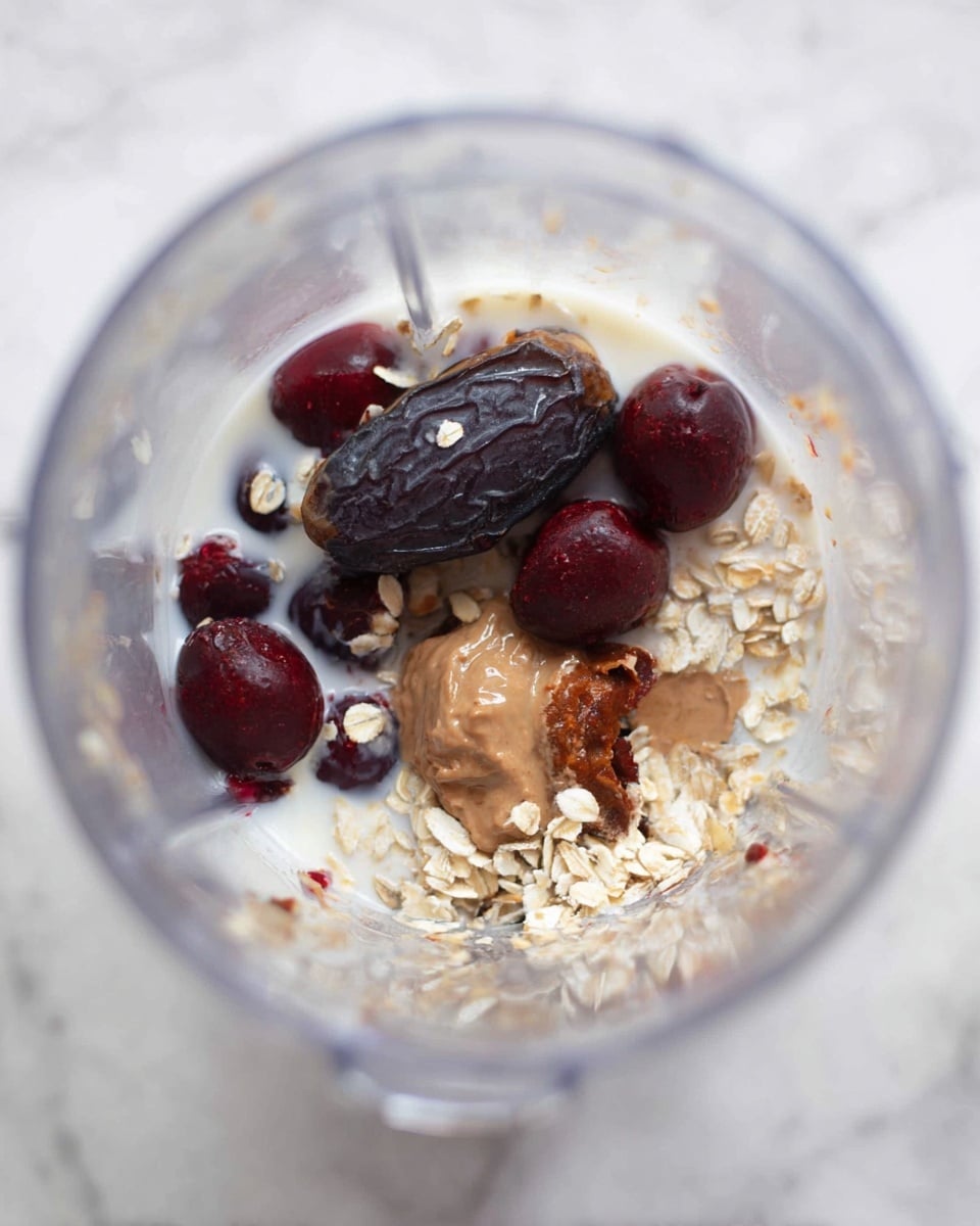 Inside a clear blender jar, there is a mix of ingredients in one layer. The bottom has light oatmeal flakes scattered around. Mixed with the oats are dark red cherries, some with brown nut butter on top, showing a smooth texture. Near the top, there is a dark, wrinkled date resting among the oats and cherries. All the ingredients sit on a thin layer of white milk at the bottom. The photo is taken from above, showing the clear jar edges against a white marbled background. photo taken with an iphone --ar 4:5 --v 7