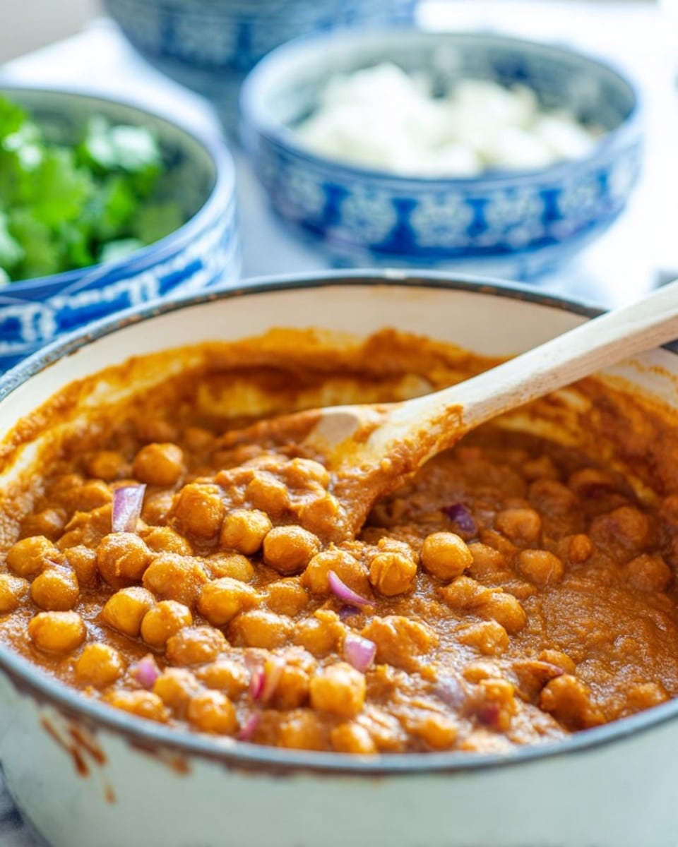 A blue pot filled with a thick orange-brown stew made of chickpeas and small pieces of vegetables, with a wooden spoon resting inside the pot. Behind the pot, there are two white bowls with blue floral patterns, each containing white cottage cheese topped with green herbs. To the left, a stack of golden flatbreads is slightly blurred, and in the background, some green herbs lay on a white marbled surface. The lighting is bright and natural. photo taken with an iphone --ar 4:5 --v 7