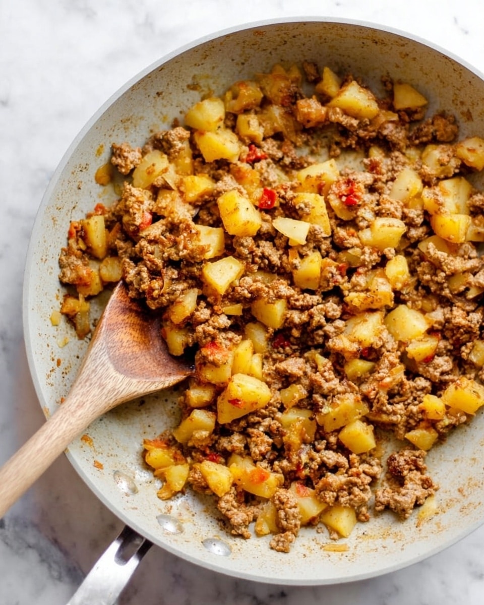 A close-up of a white frying pan filled with cooked ground meat mixed with chopped yellow potatoes and translucent cooked onion pieces. The mixture has a slightly browned texture, indicating it has been pan-fried, with some reddish bits that look like small tomato pieces or seasoning. In the middle of the pan, a wooden spoon is partly buried under the mixture, with its handle pointing upward. The pan rests on a white marbled surface, and a woman's hand is just touching the handle of the wooden spoon. photo taken with an iphone --ar 4:5 --v 7