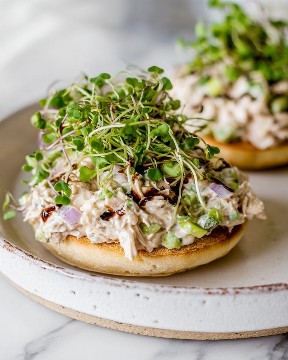 The image shows two open-faced sandwiches on a white plate with a slightly rough edge. Each sandwich has a toasted English muffin base, golden and crisp, topped with a creamy salad made of white shredded chicken mixed with light green celery pieces and small bits of purple onion. On top of the chicken salad, there is a fresh, bright green layer of microgreens with delicate stems and tiny leaves. Some dark, glossy balsamic glaze is drizzled over the microgreens, adding contrast. The plate is set on a white marbled surface, with a soft and bright background that softly blurs the second sandwich in the back. Photo taken with an iphone --ar 4:5 --v 7