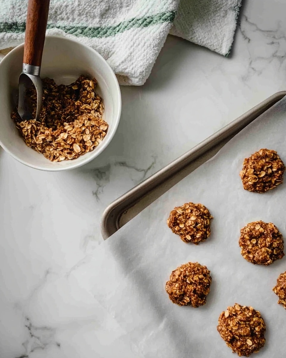 The image shows six small, round oat cookies placed on a baking sheet lined with white parchment paper. The cookies have a rough texture with visible oats and small pieces mixed in, giving them a crunchy appearance. On the left side, there is a white bowl with a wooden spoon and a metal scoop inside, containing more of the oat mixture. The background is a white marbled surface with a white and green striped cloth partially visible in the top left corner. photo taken with an iphone --ar 4:5 --v 7
