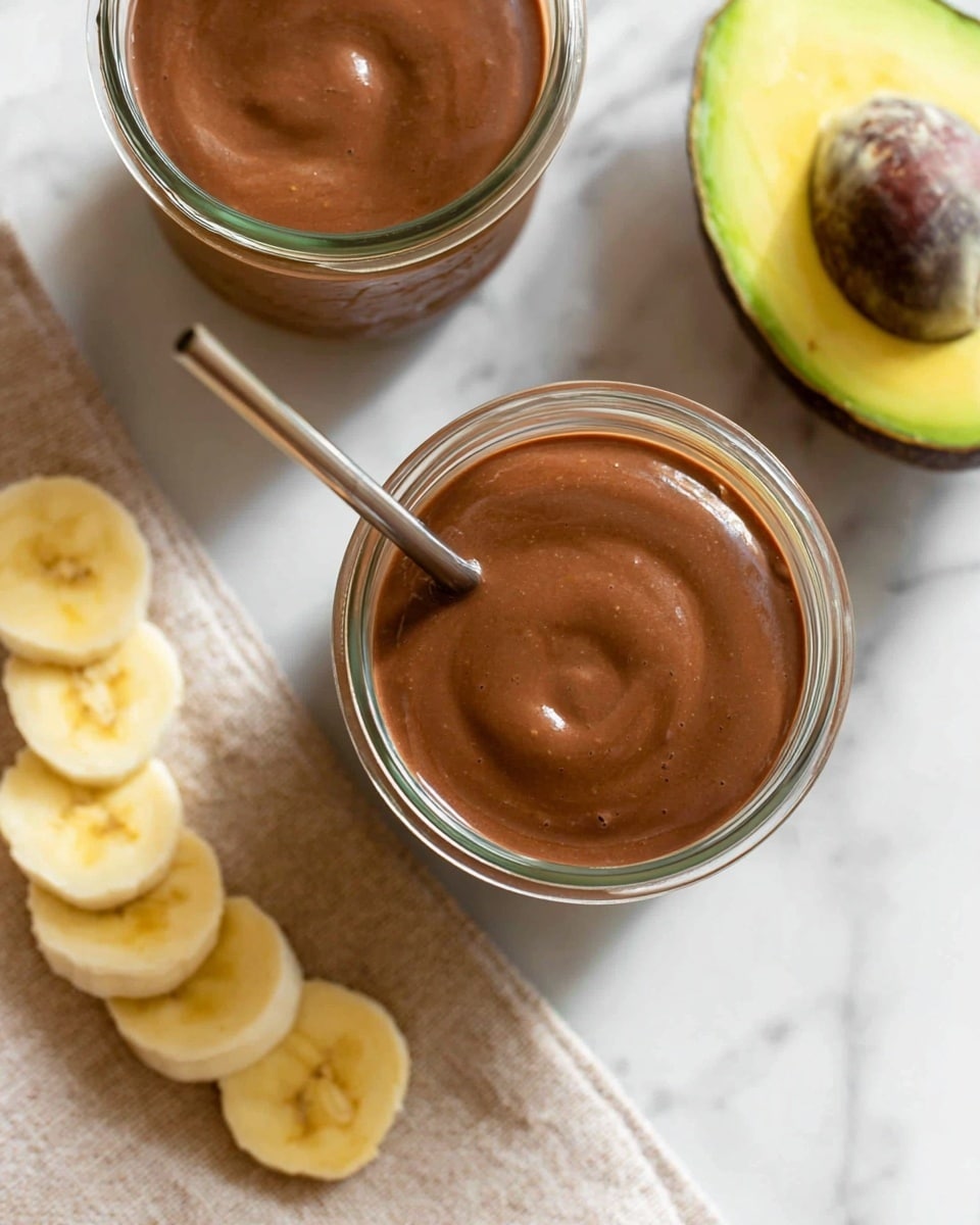 The image shows a close-up top view of two clear glass jars filled with smooth, thick brown chocolate avocado smoothie. One jar is in the foreground with a metal straw inside, and the other jar is partly visible at the top left. Around the jars, there are slices of fresh yellow banana on a beige cloth napkin at the bottom, and a halved avocado with green skin and yellow-green flesh placed on a white marbled surface to the right. The scene has a clean and fresh look. Photo taken with an iphone --ar 4:5 --v 7