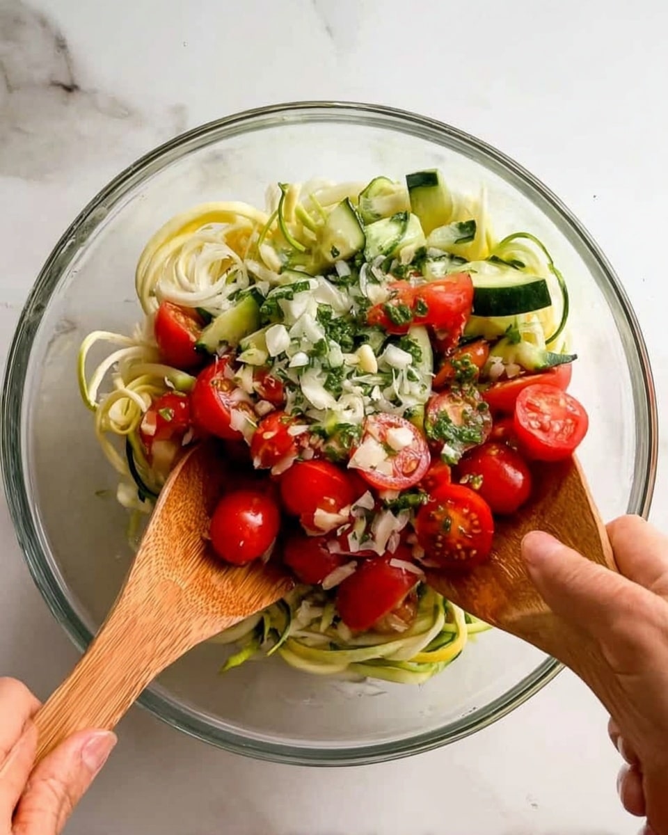 A clear glass bowl filled with a fresh vegetable salad being mixed by a woman's hand holding wooden salad utensils. The salad has a bottom layer of pale yellow spiralized zucchini noodles, topped with sliced white onions and small bright green cucumber pieces. Over this are vibrant red halved cherry tomatoes scattered around, with finely chopped green herbs and small bits of white garlic sprinkled on the top. The bowl sits on a white marbled surface. photo taken with an iphone --ar 4:5 --v 7