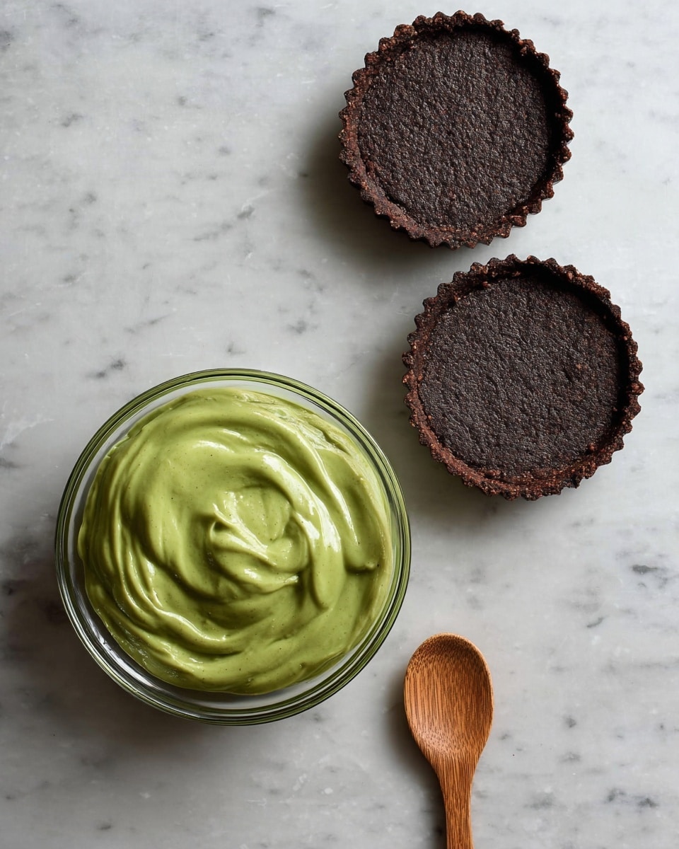The image shows two small tart crusts with a dark brown, rough texture placed at the top right and top center on a white marbled surface. Below them, there is a clear glass bowl filled with smooth, bright green creamy filling with visible swirls and peaks. To the right of the bowl, a wooden spoon with a rounded head is lying flat on the surface. The overall colors contrast between the dark crust, green filling, and light surface. photo taken with an iphone --ar 4:5 --v 7