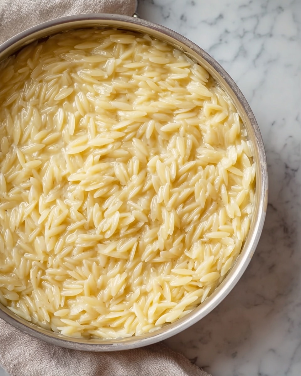 The image shows a close-up of a white bowl filled with creamy, light yellow rice. The rice looks soft and smooth, with individual grains visible and a slightly shiny, moist texture. Steam rises gently from the surface, giving a warm and fresh feel. The bowl sits on a soft beige cloth on a white marbled surface, and a light-colored wooden spoon lies next to it. The background is softly blurred with a light gray tone. photo taken with an iphone --ar 4:5 --v 7