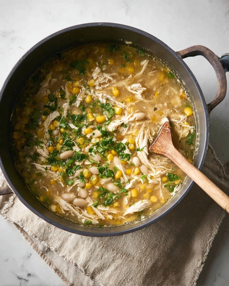 A dark gray pot filled with a soup that has multiple layers: light yellow broth at the base, with visible shredded white chicken pieces, bright yellow corn kernels, white beans, and chopped green herbs scattered on top. A wooden spoon stirs the mixture from the right side, and the pot sits on a beige cloth with a white marbled surface underneath. Photo taken with an iphone --ar 4:5 --v 7