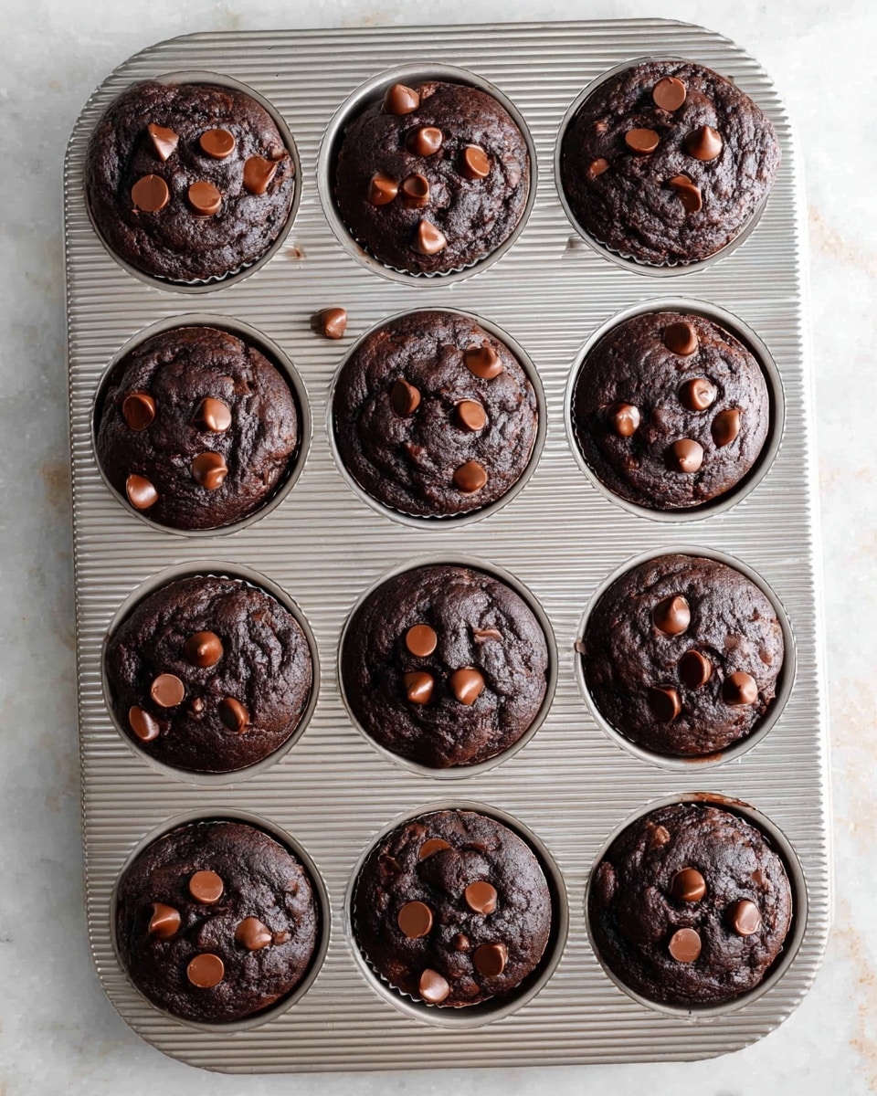 Twelve dark chocolate muffins sit in a silver metal muffin tin with a ribbed texture, each muffin topped with scattered milk chocolate chips. The muffins have a soft, slightly cracked surface indicating a rich, moist texture inside. The muffin tin rests on a white marbled surface that adds a clean, bright contrast to the deep brown color of the muffins. Photo taken with an iphone --ar 4:5 --v 7