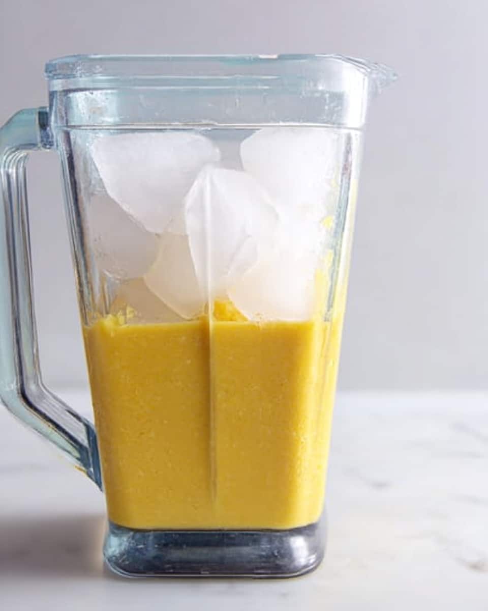 The image shows a clear blender filled with two layers of ingredients. At the bottom, there is a thick yellow mixture with a smooth texture, filling about half the blender. On top of this yellow layer, there are several large ice cubes that take up the rest of the space, creating a contrast between the solid white ice and the yellow liquid beneath. The blender is set against a white marbled background. Photo taken with an iphone --ar 4:5 --v 7