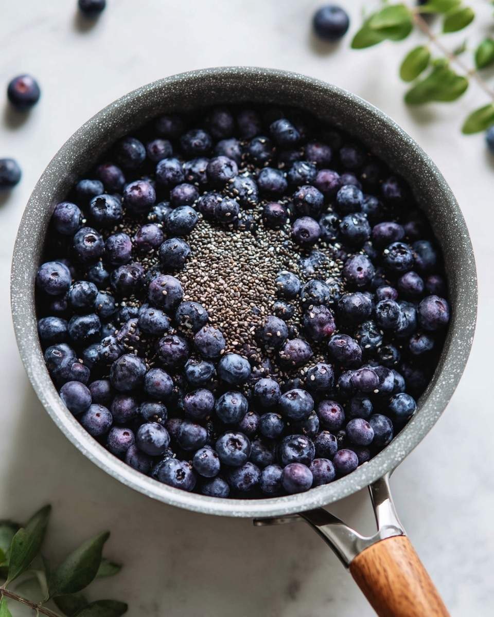 The image shows a gray pot filled with a single layer of small, dark purple blueberries spread evenly across the bottom. There is a sprinkle of tiny black and white chia seeds scattered roughly on top of the blueberries. The pot is on a white marbled textured surface with some green leaves blurred in the background. The wooden handle of the pot is visible on the right side. photo taken with an iphone --ar 4:5 --v 7