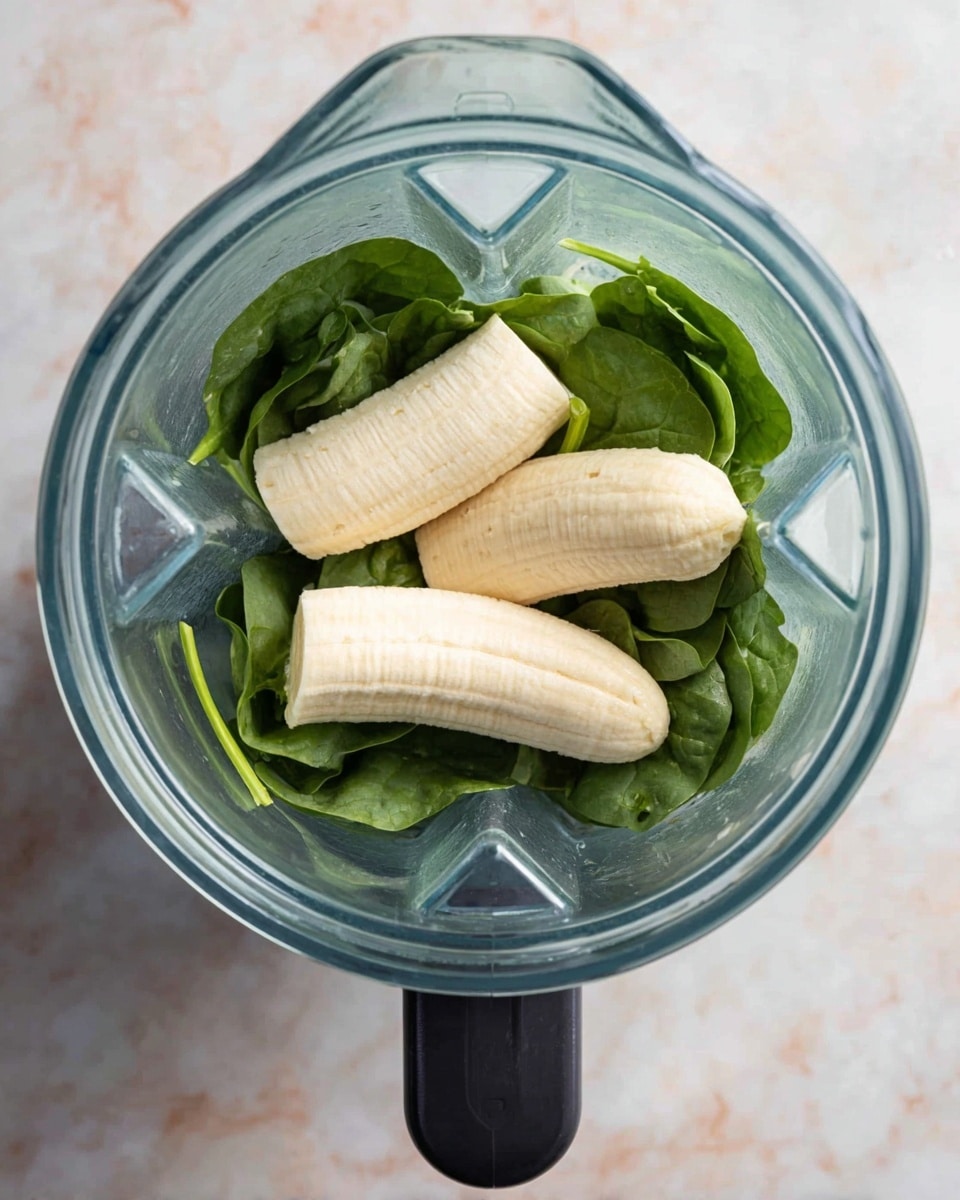 The image shows a clear blender container from above, with three peeled bananas placed on top of a layer of fresh green spinach leaves. The bananas are pale yellow with a smooth texture, and they rest in the center of the leafy spinach, which has a vibrant, slightly shiny green color with visible veins. The blender container is transparent with a black handle at the bottom. The background is a white marbled texture. photo taken with an iphone --ar 4:5 --v 7