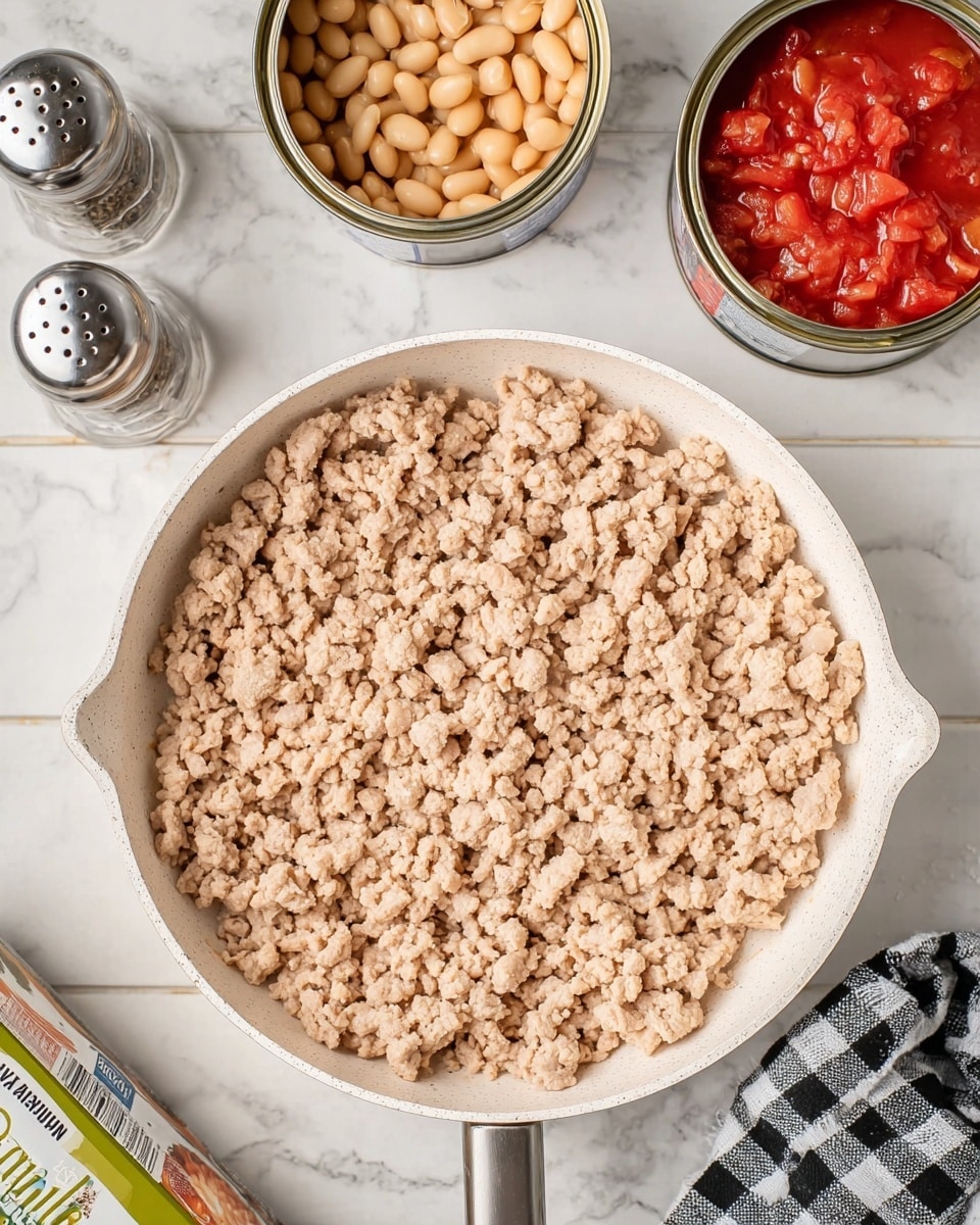 A white pan is filled with light beige cooked ground meat with a crumbly texture, spread evenly across the pan's base. To the top right of the pan, there are two open cans, one with light tan beans and the other with chunky bright red tomatoes in juice. Nearby are two metal shakers, one with pepper and the other with salt, both showing some wear and a little rust. On the lower left, there is a container with green and white packaging partially visible. The scene is set on a white marbled surface with a black and white checkered cloth in the bottom right corner. Photo taken with an iphone --ar 4:5 --v 7