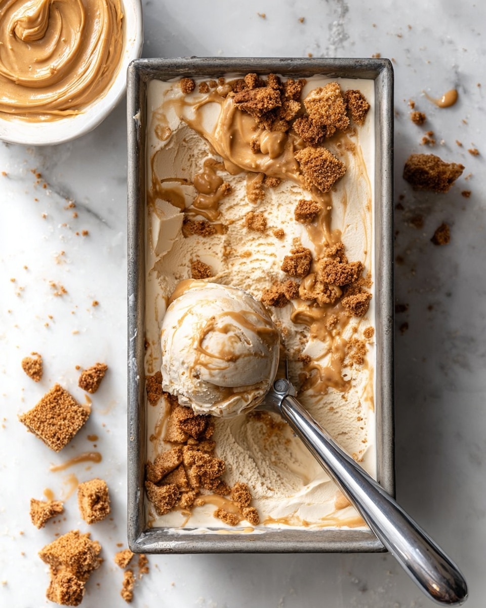 A rectangular metal container is filled with creamy beige ice cream that has swirls of caramel or peanut butter running through it. On the top surface, there are chunks of crumbly brown cookie or biscuit pieces scattered unevenly, adding texture and color contrast. A silver ice cream scoop is scooping out a round ball from the middle, showing the creamy inside mixed with the cookie bits. To the left side, on a white marbled surface, a white bowl holds more smooth caramel or peanut butter spread with a swirl pattern on top. There are small brown crumb pieces around the container on the surface. The overall scene is bright with soft light and has a clean, minimal look. photo taken with an iphone --ar 4:5 --v 7