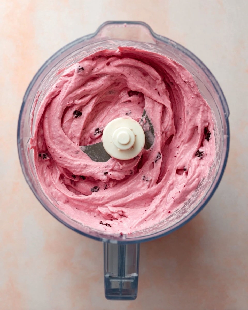 The image shows a close-up top view of a clear blender jar filled with thick, creamy, pink mixture that has small darker pieces mixed inside. The creamy texture has swirls and soft peaks, with a visible white blade in the center of the blender. The blender is placed on a white marbled surface. photo taken with an iphone --ar 4:5 --v 7