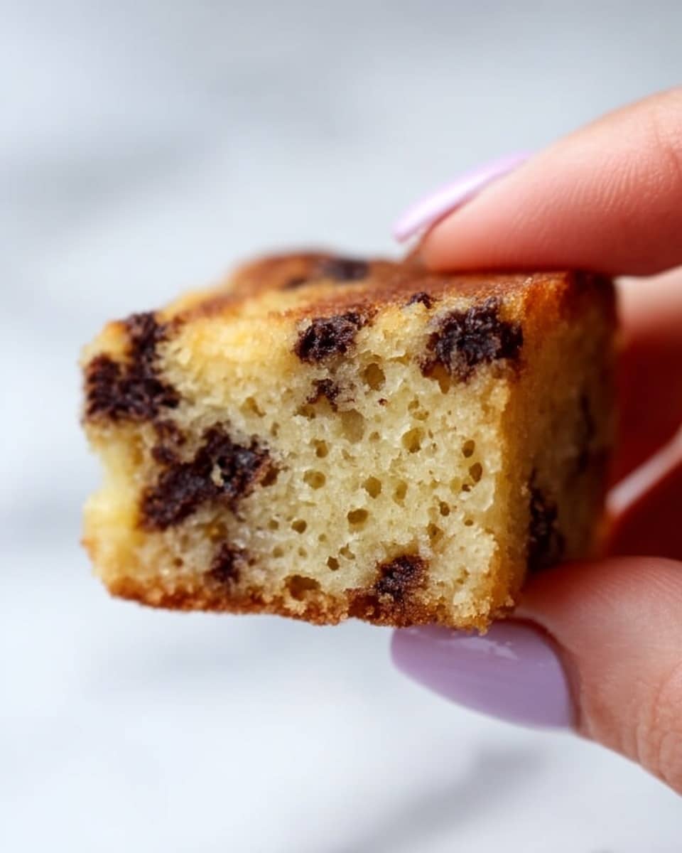 A close-up view of a small, square piece of light golden cake with visible dark chocolate chips scattered inside. The cake has a soft and moist texture with tiny air bubbles, and the edges look slightly crispy. A woman's hand with light purple nail polish is holding the cake piece gently between thumb and forefinger. The background is a white marbled texture. photo taken with an iphone --ar 4:5 --v 7