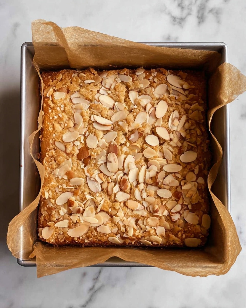 A square baked cake in a silver baking pan lined with brown parchment paper, topped with a generous layer of sliced almonds scattered evenly across the golden-brown surface, showing a slightly rough and crumbly texture. The cake's edges are slightly darker, indicating a baked crust. The background is a white marbled texture. photo taken with an iphone --ar 4:5 --v 7