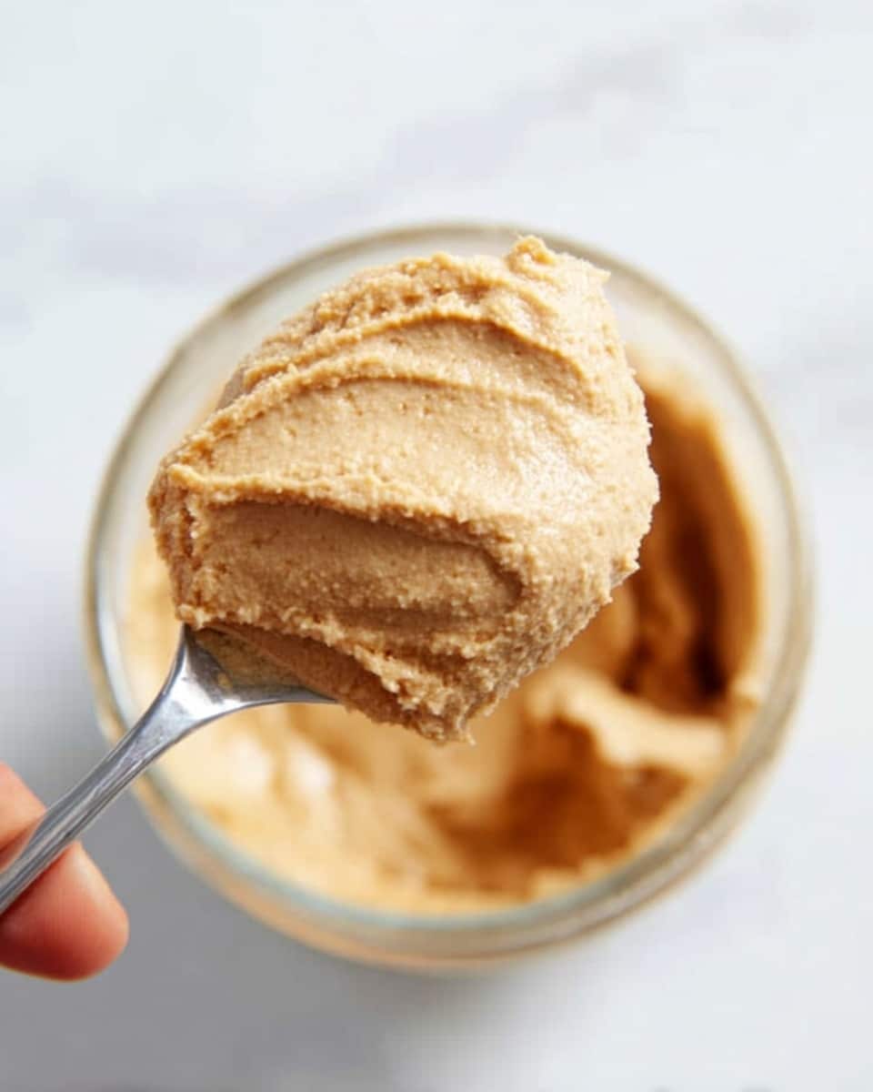 The image shows a close-up of a spoon holding a thick and creamy pale brown mixture with a slightly rough texture. The spoon is positioned above a clear glass container that has some of the same mixture inside. The background is a soft white marbled surface, giving a clean look. A woman's hand is gently holding the spoon. The photo is taken with an iphone --ar 4:5 --v 7
