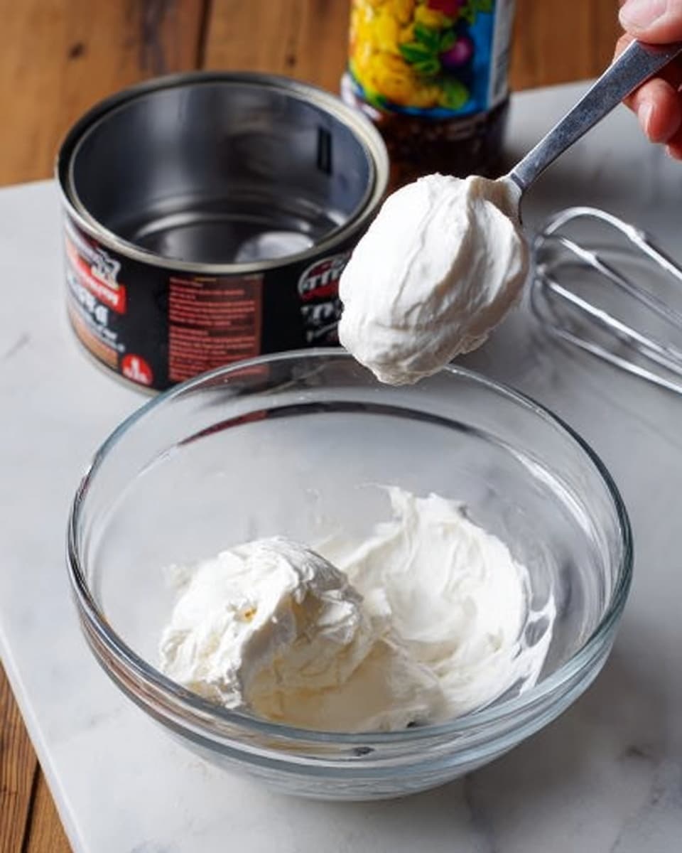 The image shows a clear glass bowl on a white marbled surface, containing two smooth, thick white cream scoops. Above the bowl, a woman's hand holding a spoon is lifting more white cream from an open can with a black and red label, positioned behind the bowl. To the right, there is a silver metal whisk resting on the surface, and to the left, partially visible bottles with colorful labels. The scene is well-lit, focusing on the creamy texture and the clear glass bowl. photo taken with an iphone --ar 4:5 --v 7