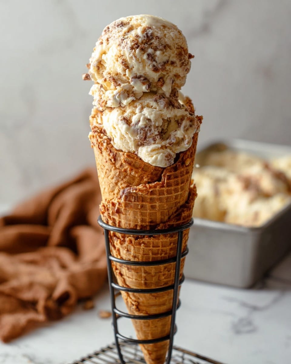 A tall stack of seven golden brown waffle cones is held upright by a black metal cone holder, with two scoops of ice cream on top. The ice cream is creamy off-white with swirls and chunks of light and dark brown mixed throughout, creating a textured and rich look. The stack is set against a white marbled surface with a soft brown cloth in the background and a metal container with more ice cream blurred out behind. Photo taken with an iphone --ar 4:5 --v 7