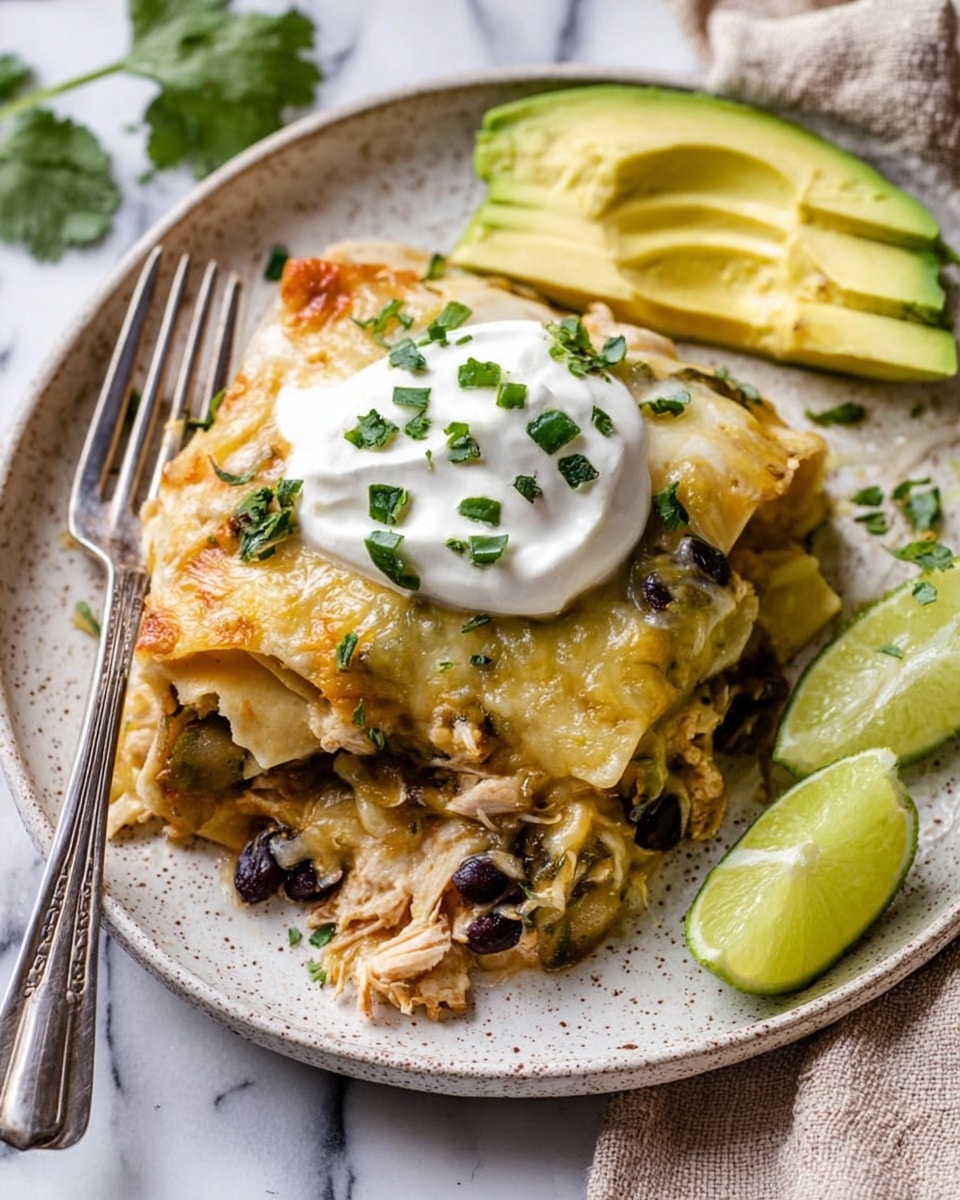 A white speckled plate holds a layered dish starting with a base of soft, light yellow pasta sheets mixed with shredded light-colored chicken and dark black beans. The next layer is melted pale yellow cheese covering the pasta and chicken, sprinkled with chopped green herbs. On top sits a dollop of smooth white sour cream with green herb pieces scattered around it. Two thick slices of bright yellow-green avocado rest on the right edge, and two thin lime wedges with a fresh green rind stand at the back of the plate. A silver fork lies on the left side of the plate. The plate is on a white marbled surface with a beige cloth in the background. photo taken with an iphone --ar 4:5 --v 7