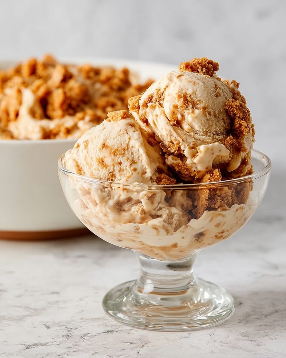 A clear glass dessert cup holds two large scoops of creamy ice cream with a light tan color, speckled and swirled with thick caramel or brown crumbs on top and mixed throughout, creating a textured look. The ice cream appears soft and rich, with uneven clumps of crunchy cookie or crumble bits embedded in the scoops. Behind the cup, there is a white shallow bowl filled with more of the same textured ice cream, placed on a white marbled surface. The focus is on the ice cream with soft lighting, showing detailed textures and colors. photo taken with an iphone --ar 4:5 --v 7