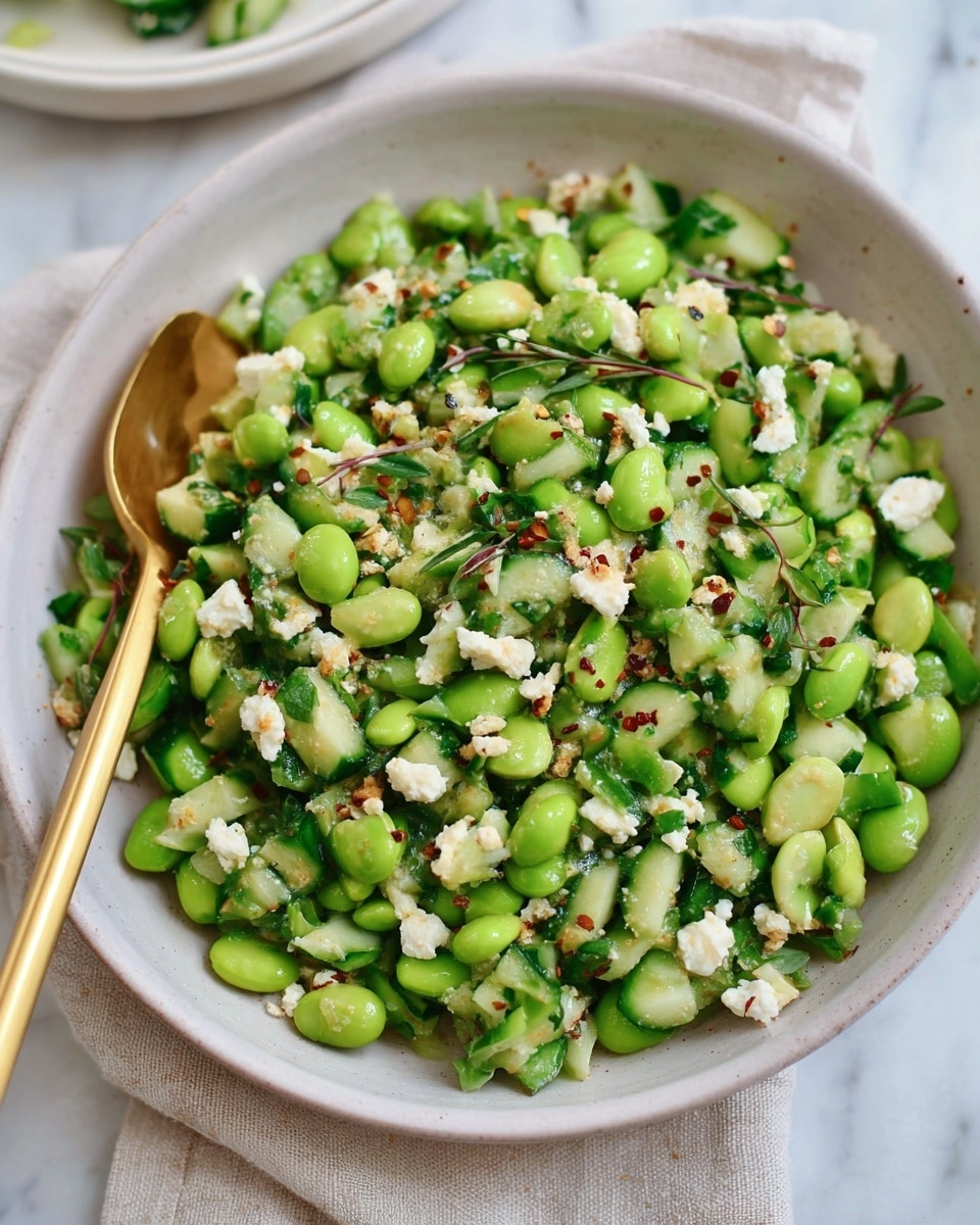 A white bowl filled with a fresh green salad that has three main parts: bright green edamame beans, chopped green cucumbers in small pieces, and small bits of white cheese scattered evenly throughout. The salad is mixed with tiny green herbs and some small red pepper flakes, giving it a colorful and fresh look. A gold spoon rests inside the bowl on the left side, the bowl sits on a light beige cloth over a white marbled surface. The colors are fresh and natural, showing a healthy and simple dish. Photo taken with an iphone --ar 4:5 --v 7