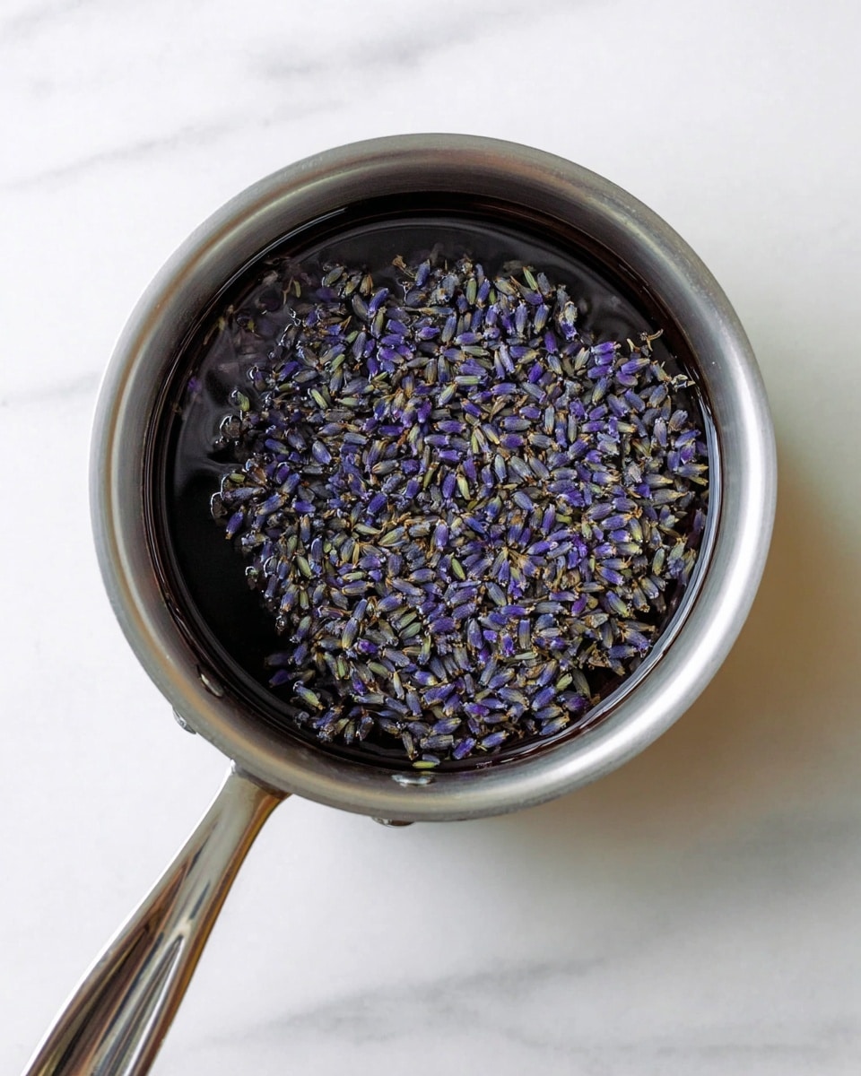 A silver metal saucepan with a long handle is shown from above, filled with water and small dried lavender flowers floating evenly on the surface. The lavender flowers are purple with bits of green stems mixed in, creating a speckled texture over the dark water. The saucepan sits on a white marbled surface that has soft gray veins running through it, adding a clean and bright background. photo taken with an iphone --ar 4:5 --v 7