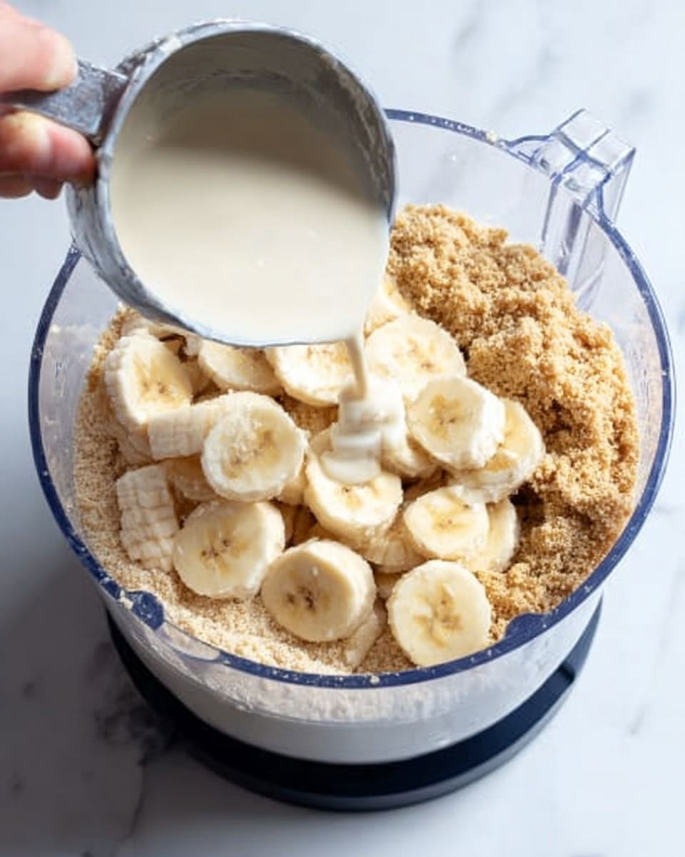 A clear food processor bowl is filled with several layers: the bottom layer consists of light brown crumbly powder, mostly covering the base; on top of that are sliced banana pieces, pale yellow with a soft texture, spread evenly; a woman's hand is pouring a smooth white liquid from a metal measuring cup into the bowl. The whole setup is on a white marbled surface. photo taken with an iphone --ar 4:5 --v 7