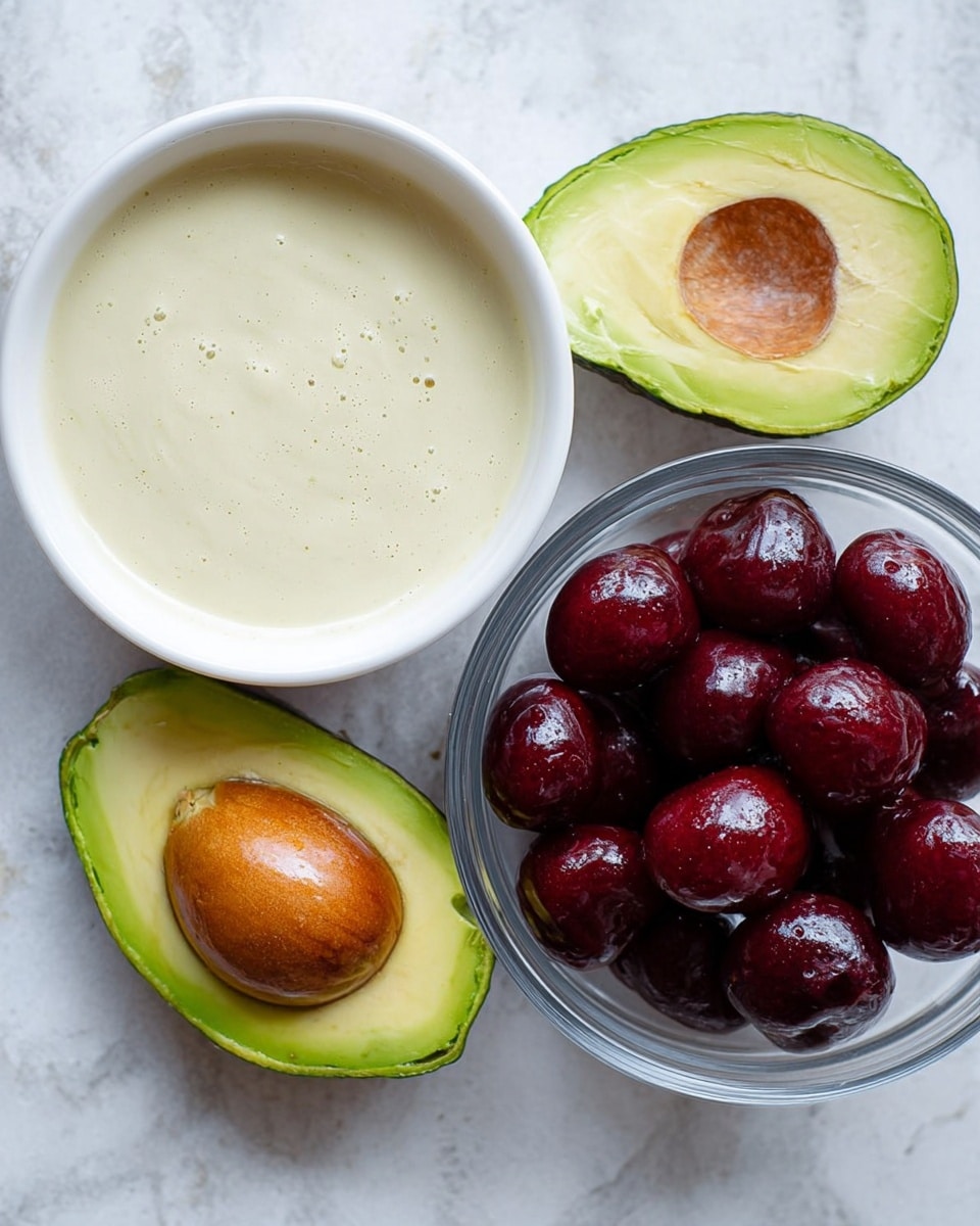 The image shows two avocado halves on a white marbled surface, one with the brown seed inside and the other with the seed removed, showing a smooth green and yellow flesh. Next to the avocados, there is a white bowl filled with a light, creamy mixture that looks smooth with tiny bubbles on the surface. Below the bowl, there is a clear glass bowl filled with shiny, dark red cherries, some of which have a slightly reflective surface, making them look fresh and juicy. Photo taken with an iphone --ar 4:5 --v 7