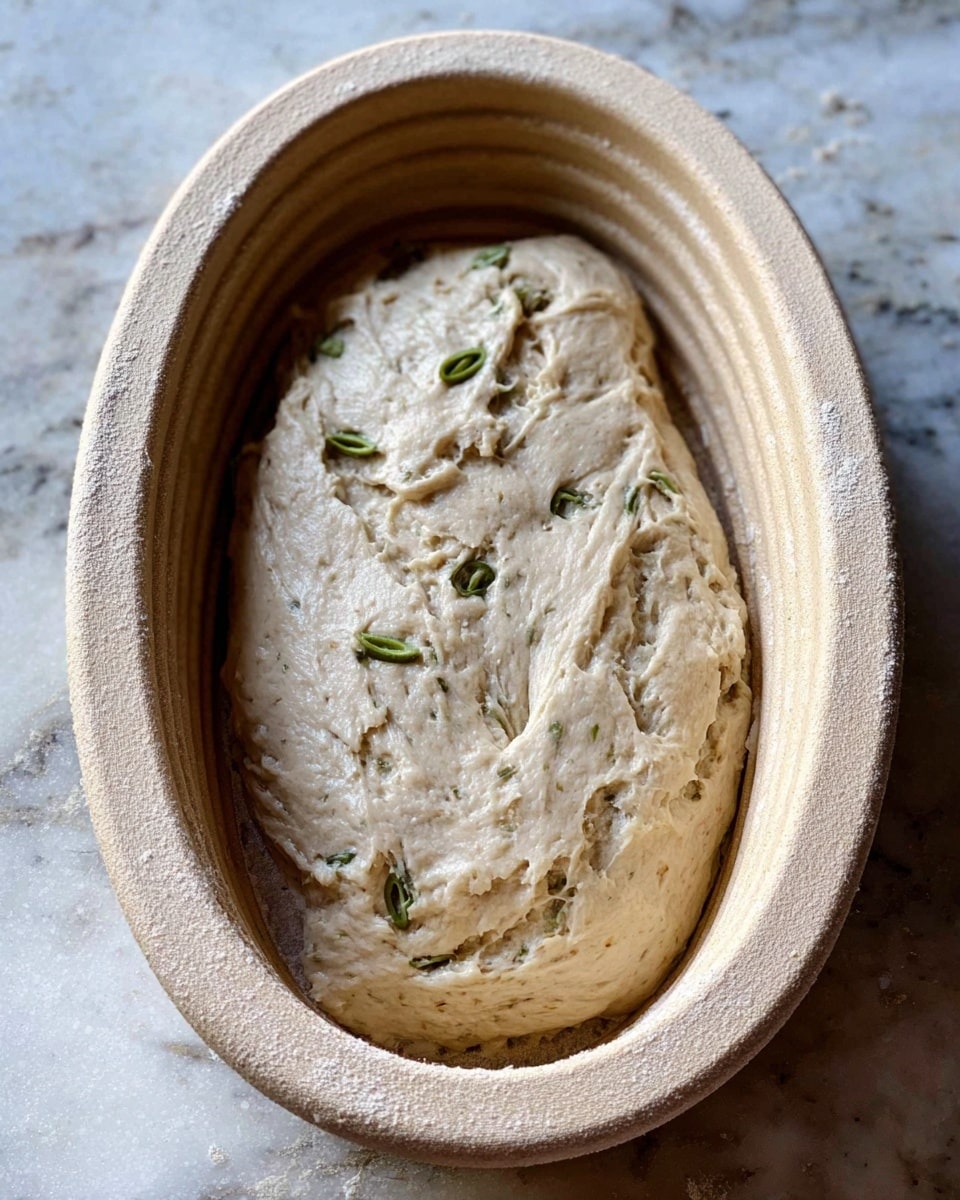 An oval-shaped raw dough rests inside a light beige oval banneton basket with grooves on the sides. The dough is pale beige with a soft, slightly sticky texture, and several small green olive pieces are embedded on the surface. The basket and dough are placed on a white marbled countertop, showing subtle veins and natural patterns. Photo taken with an iphone --ar 4:5 --v 7