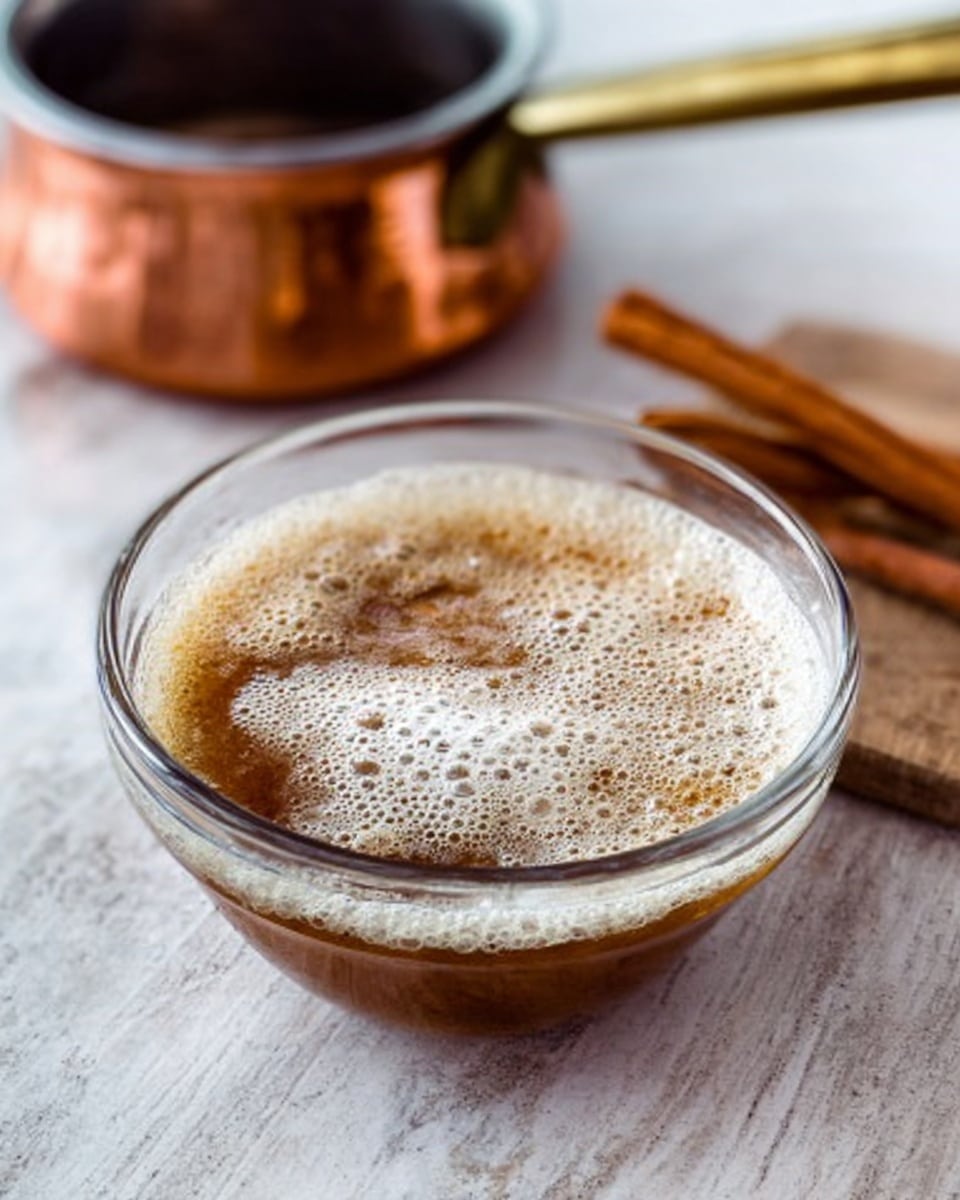 A clear glass bowl contains a layer of light brown liquid with white foam bubbles covering the top surface unevenly. Near the bowl, there are two cinnamon sticks placed side by side on the white marbled surface. Behind the bowl, a small shiny copper pot with a handle is slightly blurred but still visible. The entire scene has soft, natural light with focus on the bowl and its contents. Photo taken with an iphone --ar 4:5 --v 7