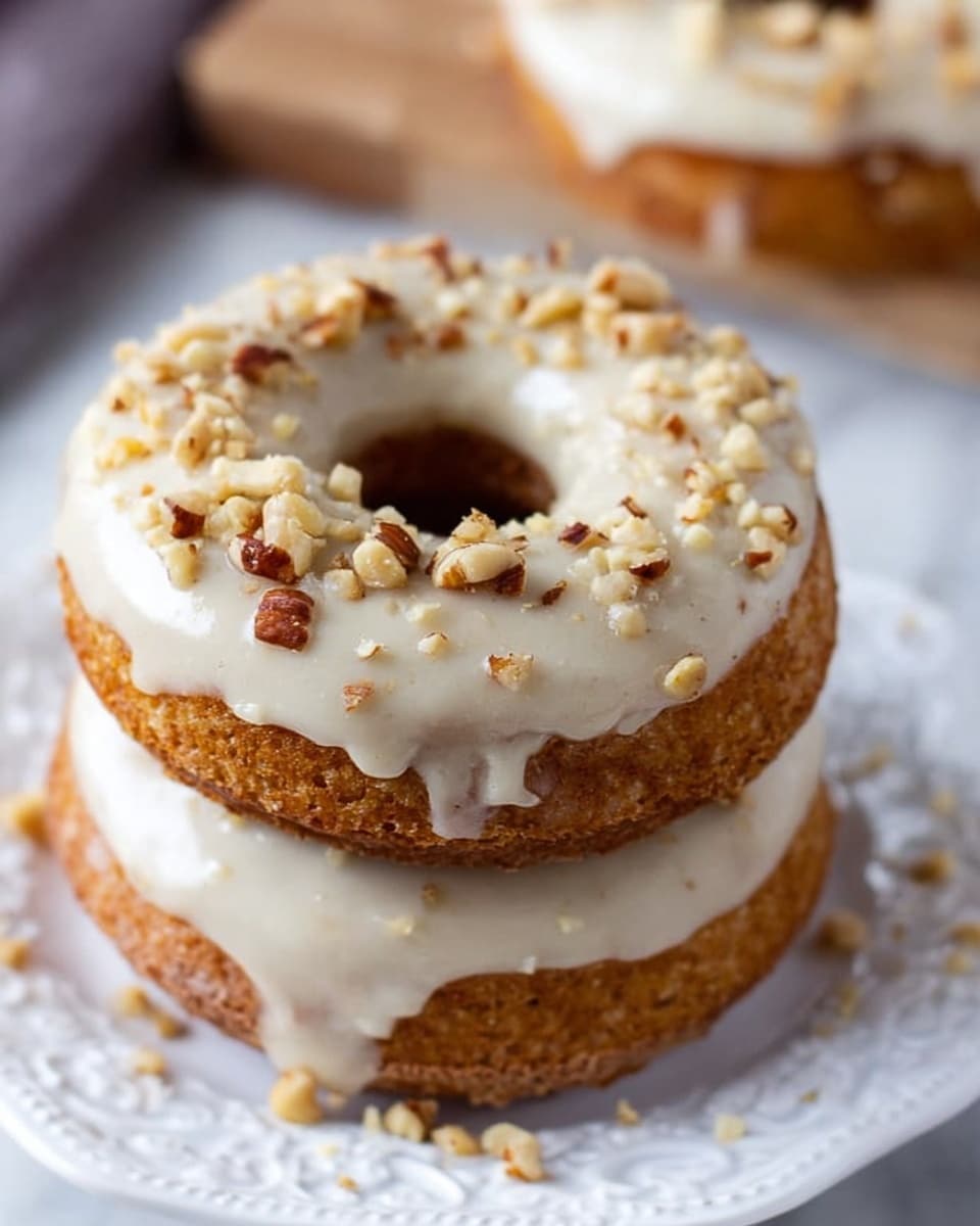 The image shows a stack of two light-brown donuts on a white plate with delicate patterned edges, placed on a white marbled surface. Each donut has a thick, smooth layer of creamy white icing on top, slightly dripping down the sides. Small pieces of crushed nuts are sprinkled evenly on the icing, adding texture and contrast. The warm tones of the donuts and nuts stand out against the white icing and plate. Part of another donut is visible blurred in the background. Photo taken with an iphone --ar 4:5 --v 7